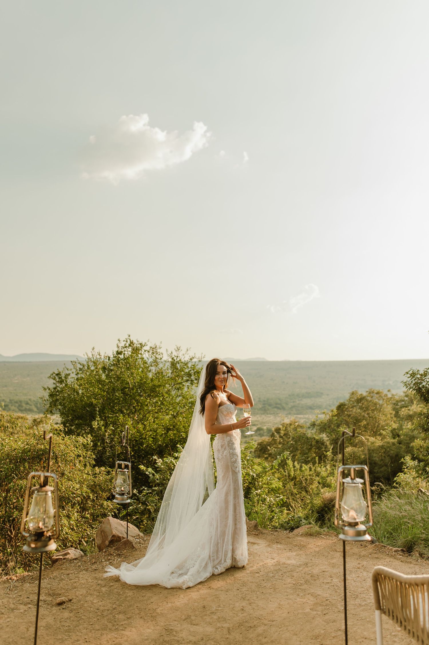 Bride in lace gown and cathedral veil stands on scenic overlook surrounded by lanterns at outdoor wedding.