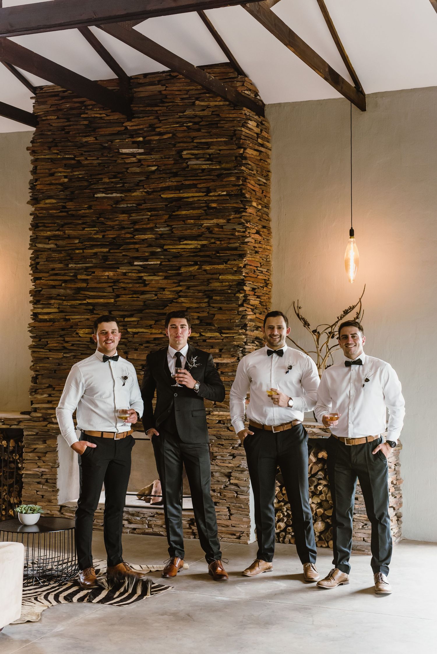 Groomsmen in formal attire pose playfully in sequence against a stone wall backdrop.