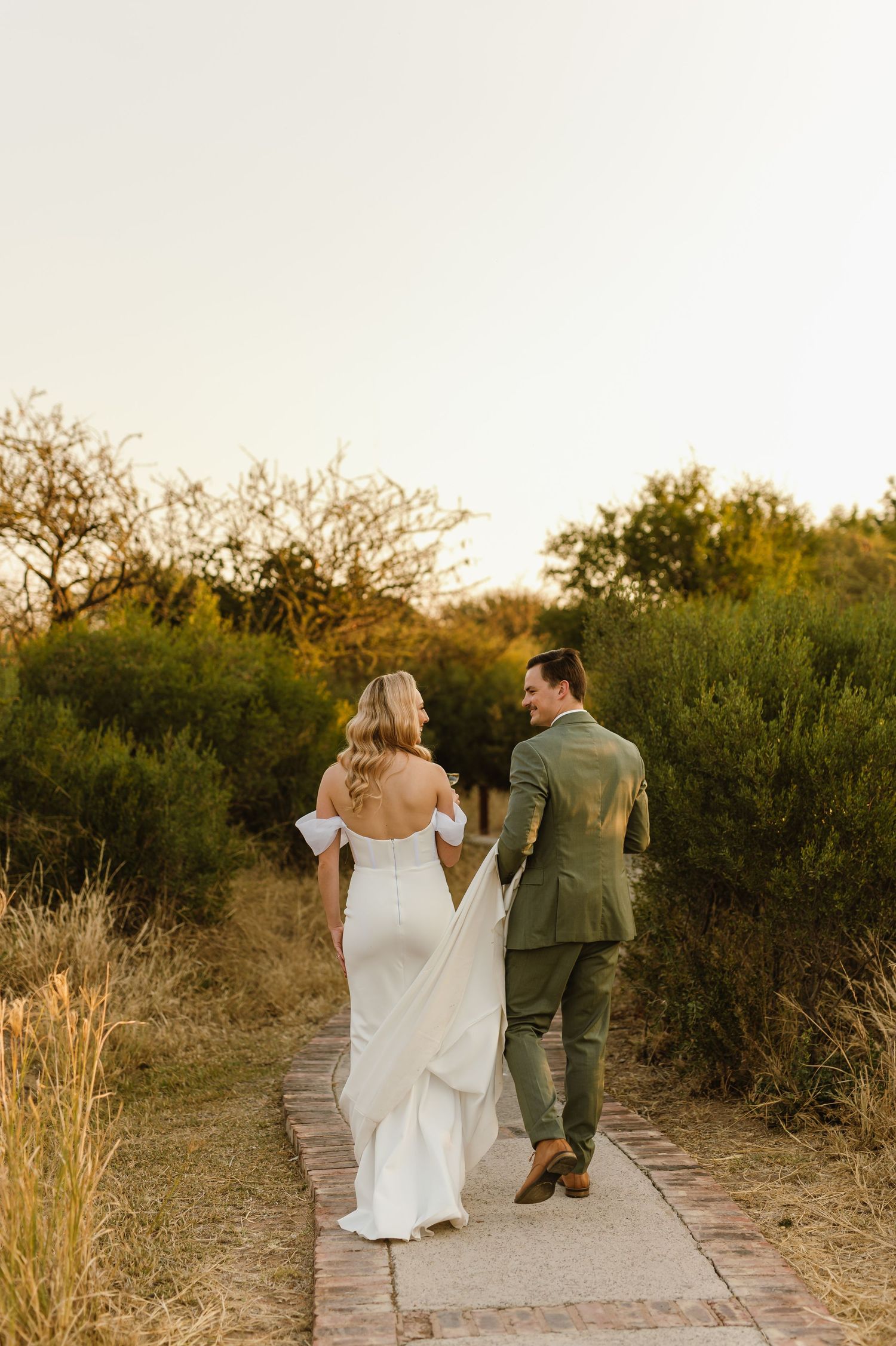 Newlyweds walk hand in hand down a sunlit path surrounded by golden grass and greenery at sunset.