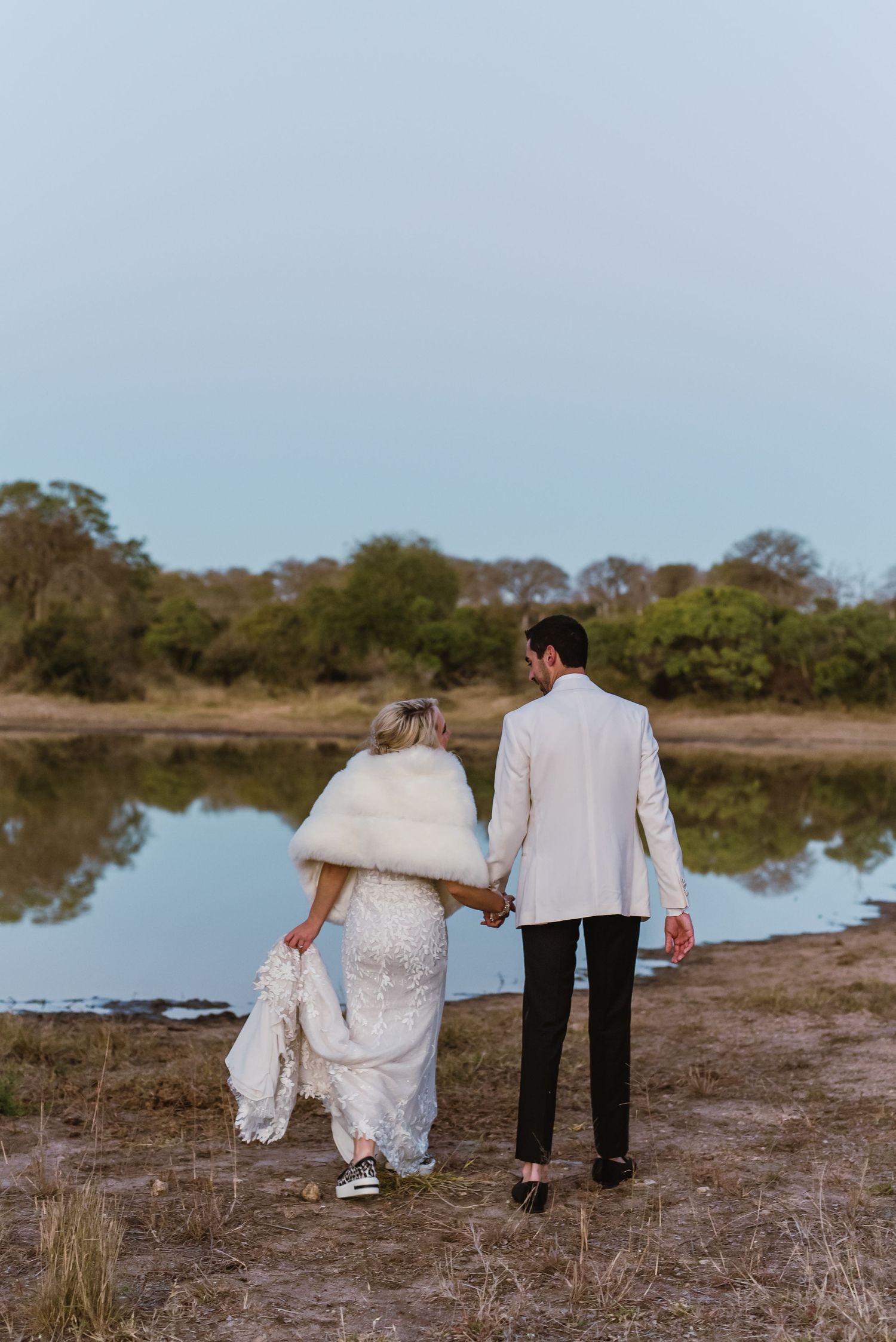 A couple in wedding attire walks along a waterfront path during sunset.