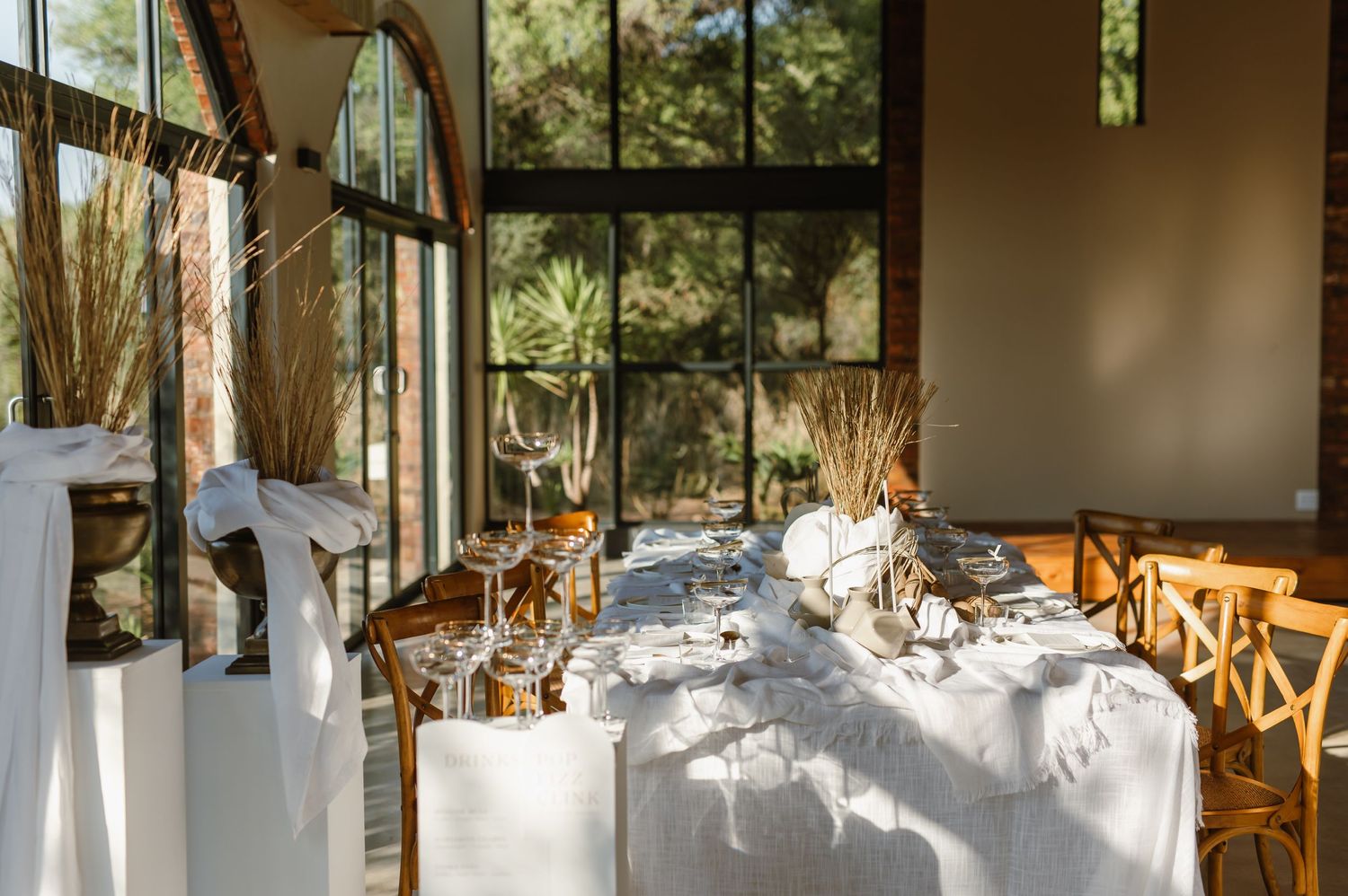 Wedding venue setup with white linens and wooden cross-back chairs in natural lighting.