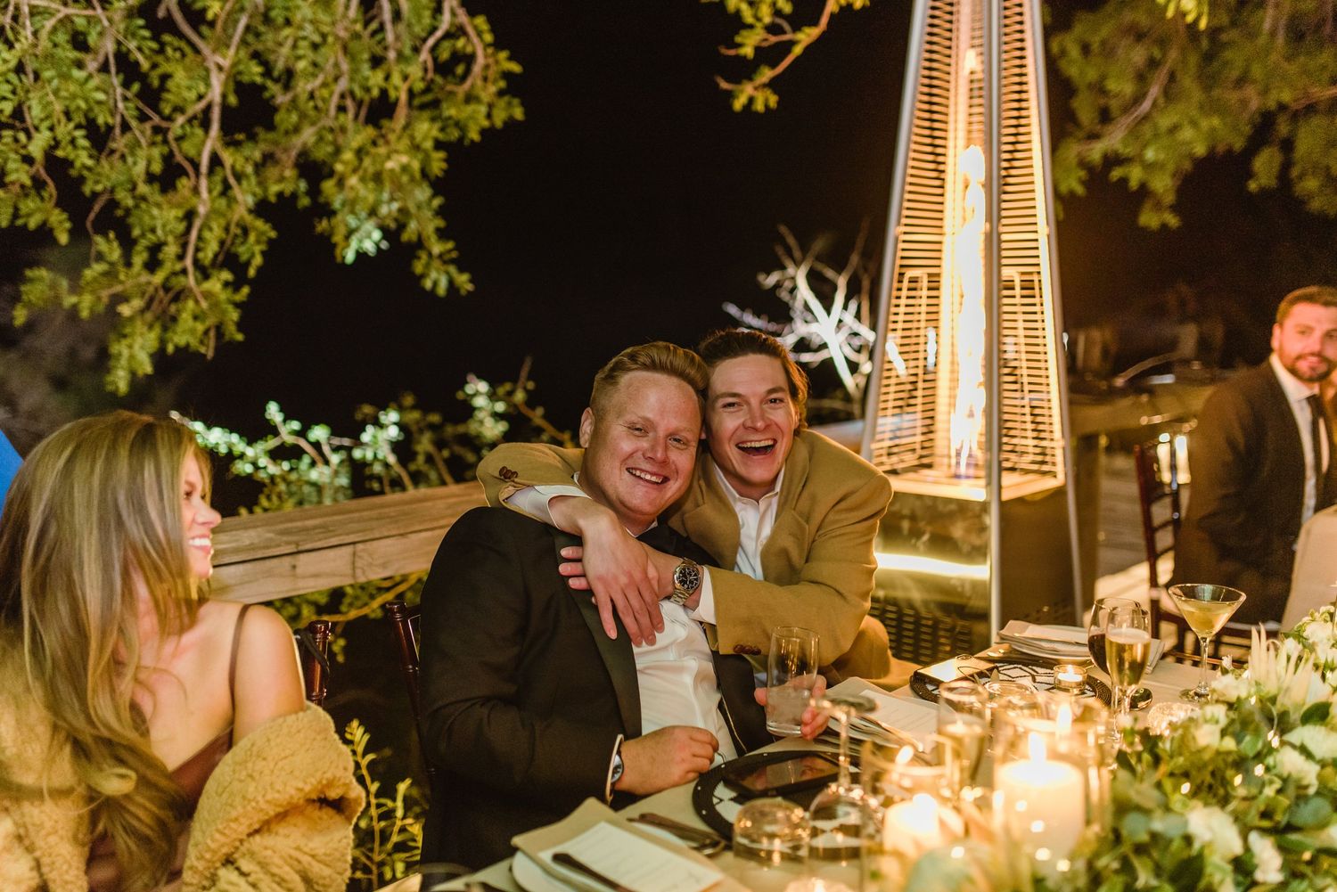 Wedding guests socializing at outdoor tables illuminated by warm candlelight and string lights.