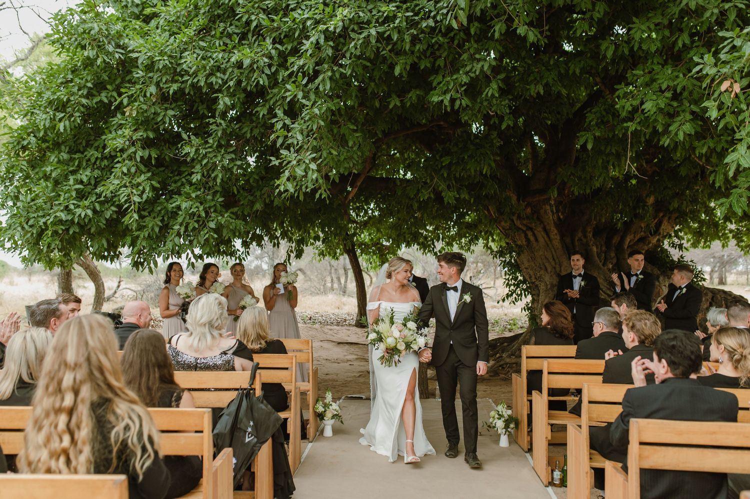 Rustic outdoor wedding venue with wooden bench seating arranged under green trees.