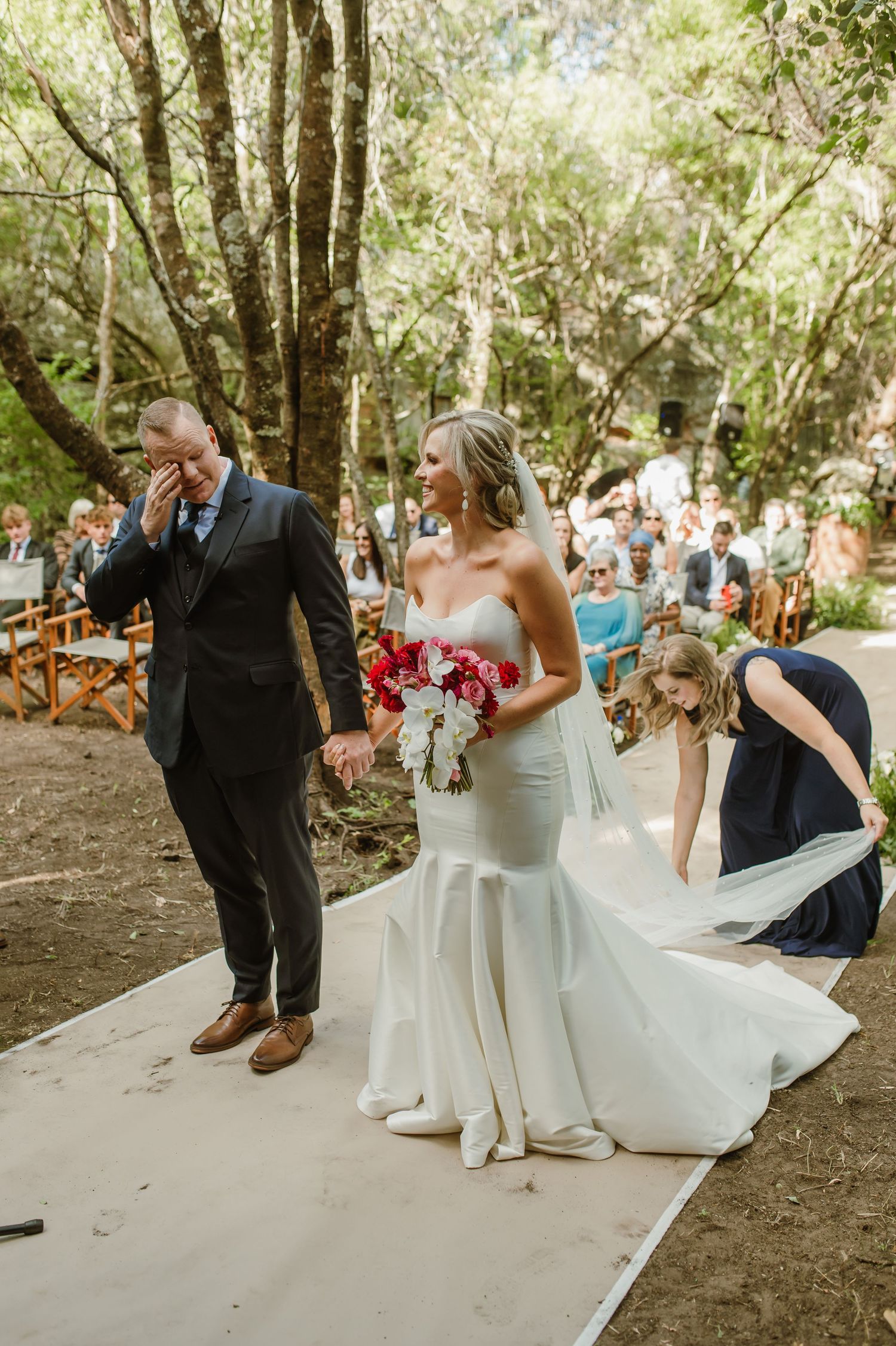 Couple exits down white aisle runner through wooded ceremony space with guests looking on.