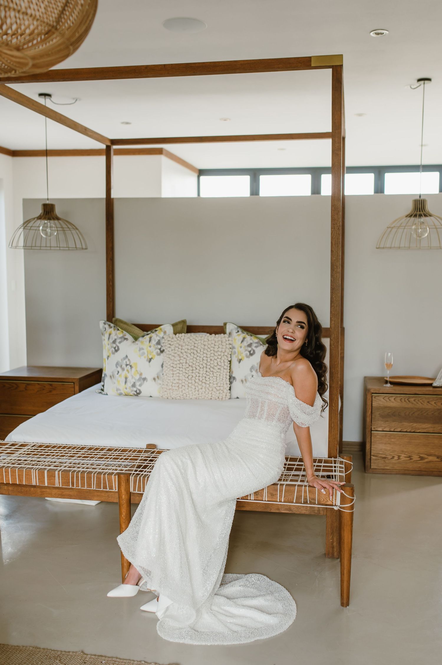 A bride in a flowing white gown poses playfully on a wooden four-poster bed in a bright modern room.