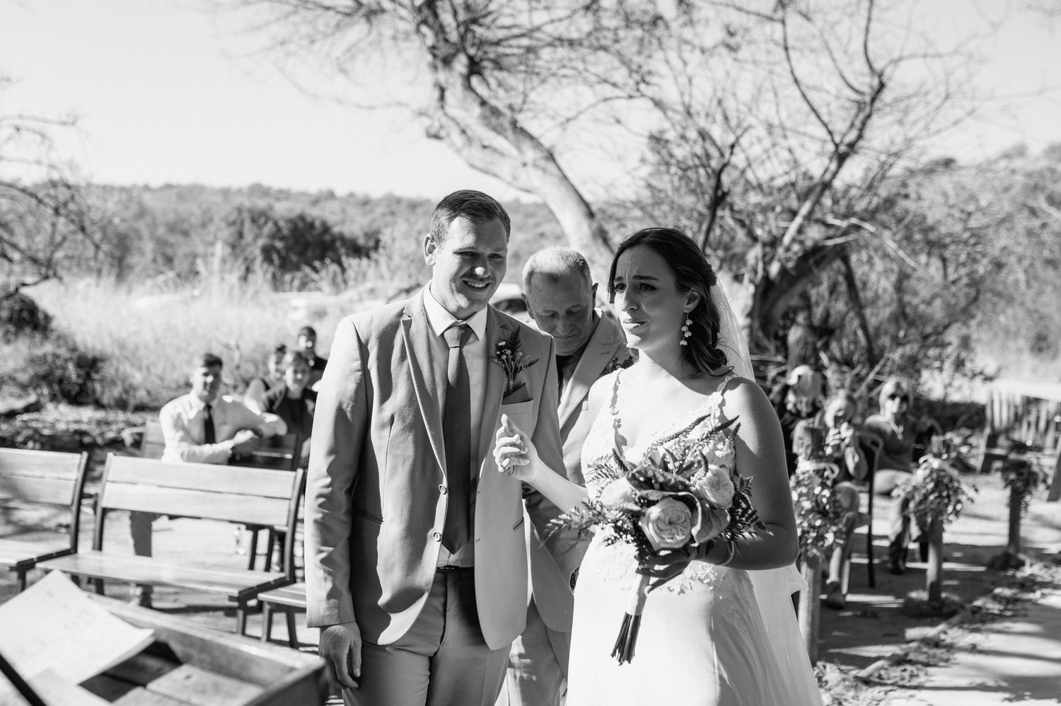 Outdoor wedding ceremony under a large tree with guests seated on wooden benches in a natural desert setting.