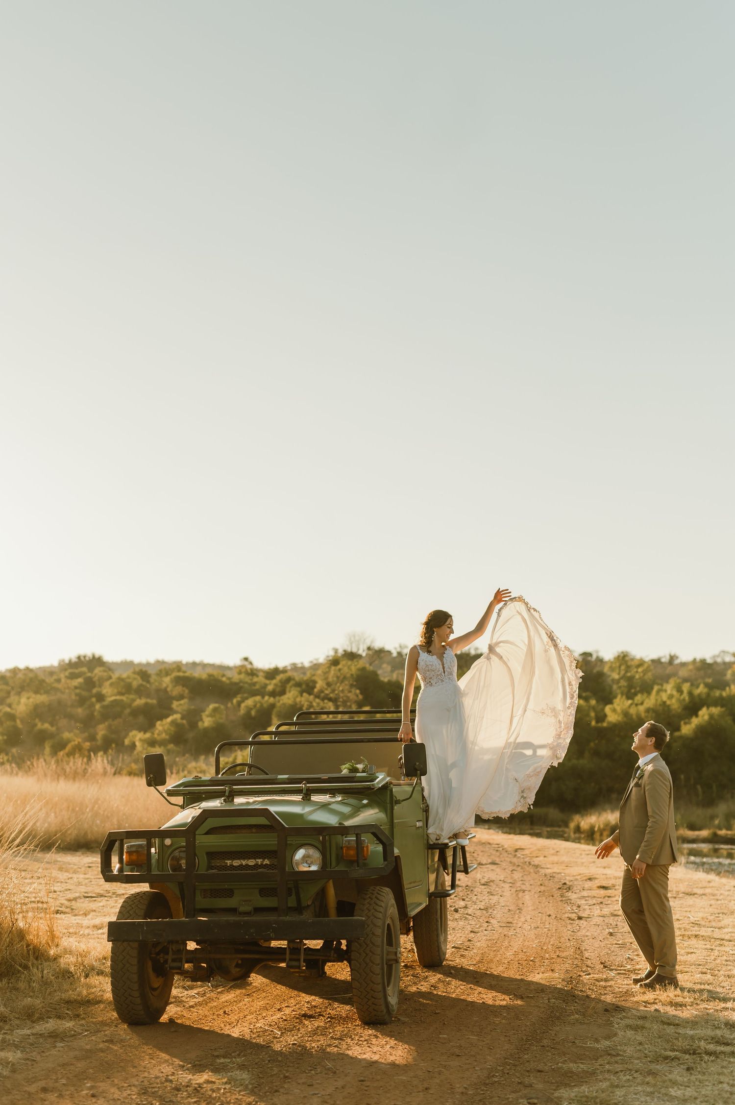 Bride stands on vintage off-road vehicle for dramatic wedding photo in rustic setting.