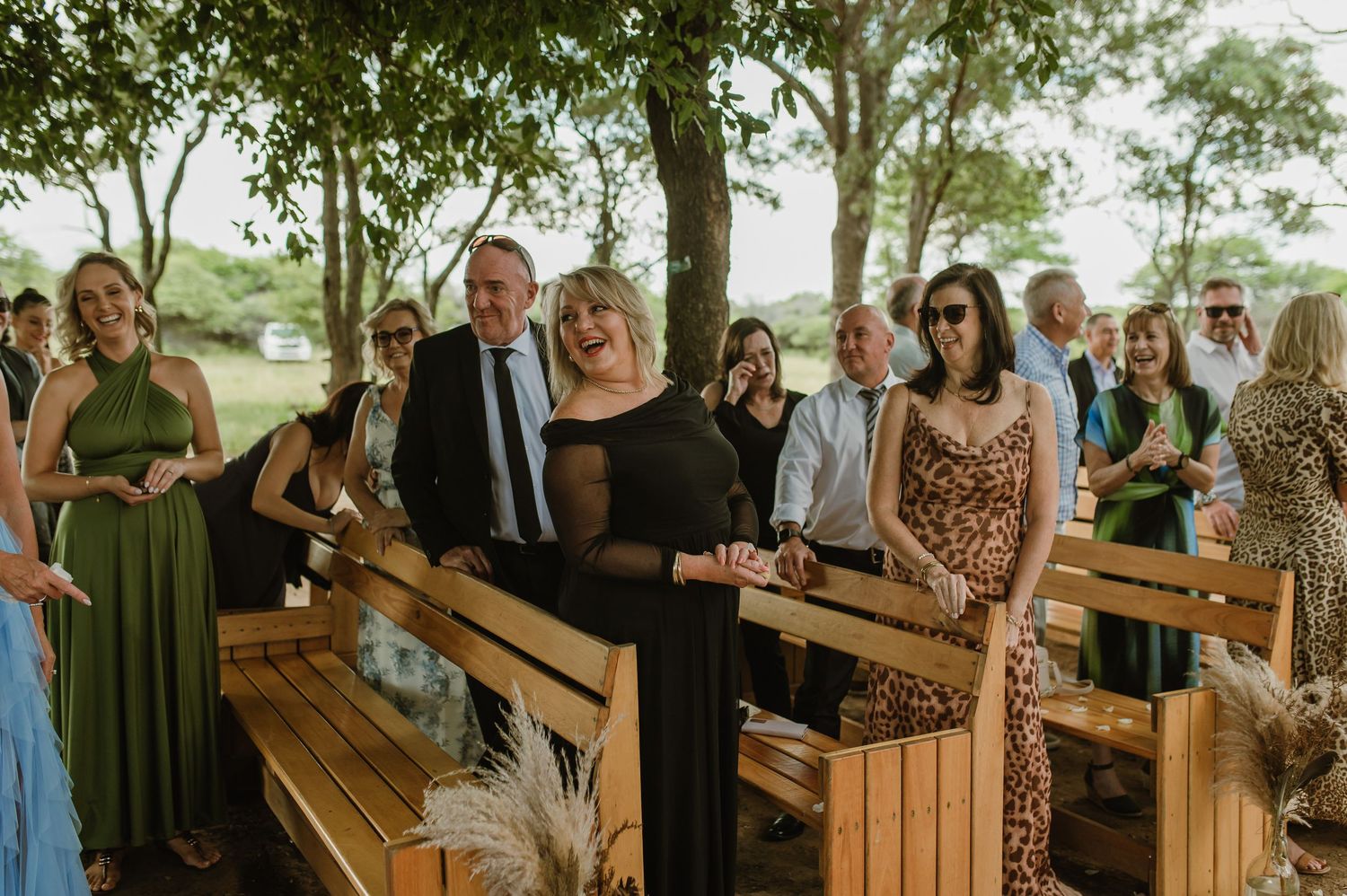 Wedding guests gathered on wooden benches at an outdoor ceremony under trees.