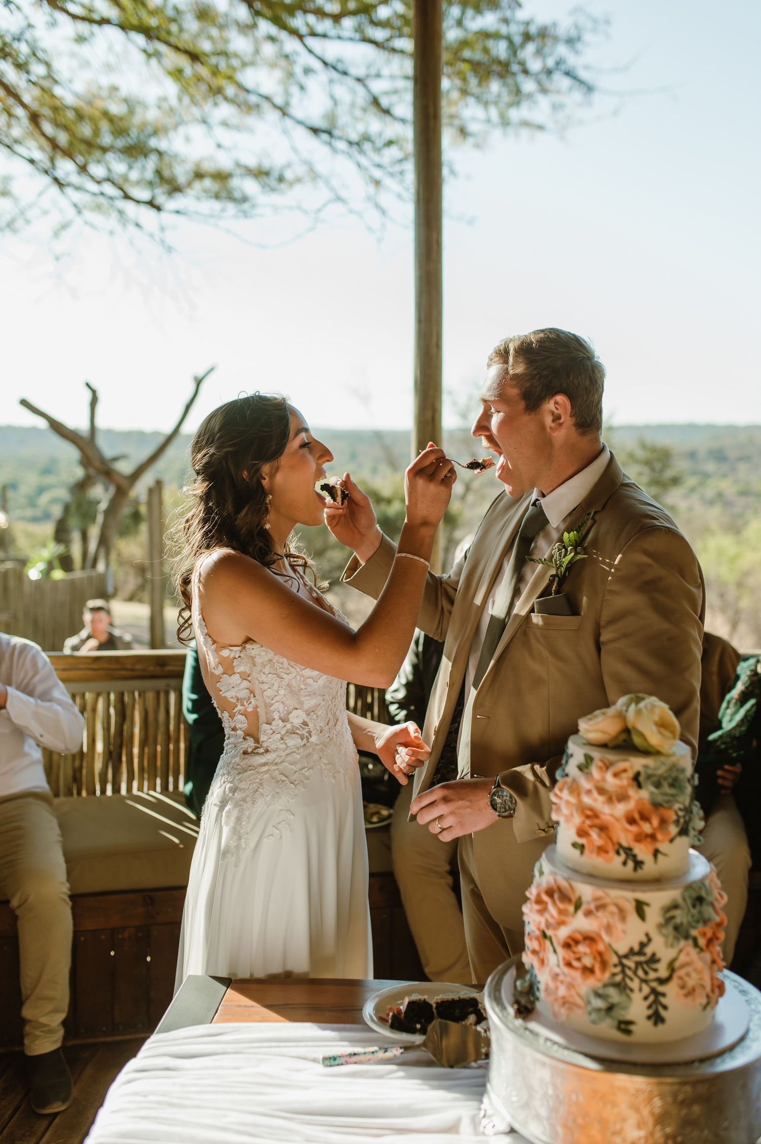 Couple sharing playful moment while feeding each other wedding cake at rustic outdoor venue.