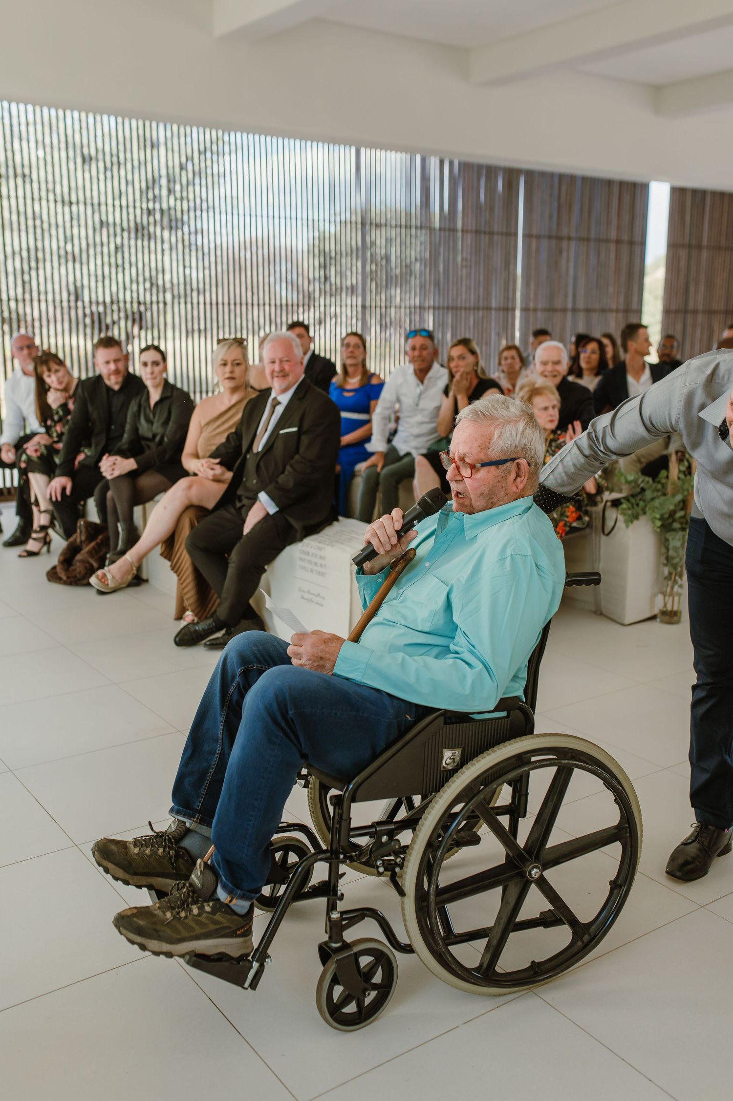 Wedding guests including elderly person in wheelchair watching ceremony in modern white venue.