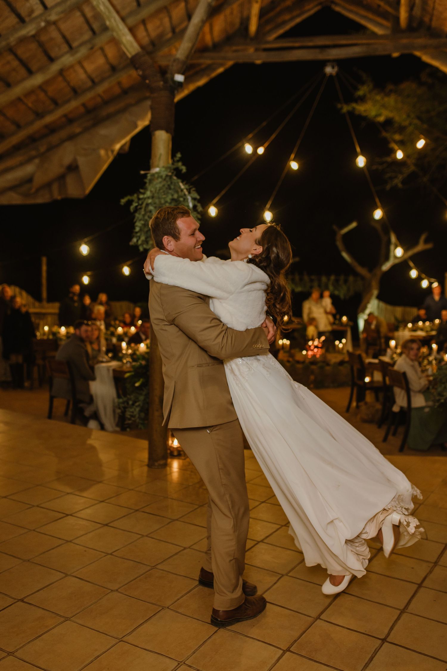 A couple dancing and spinning on a tiled floor during their wedding reception under string lights.