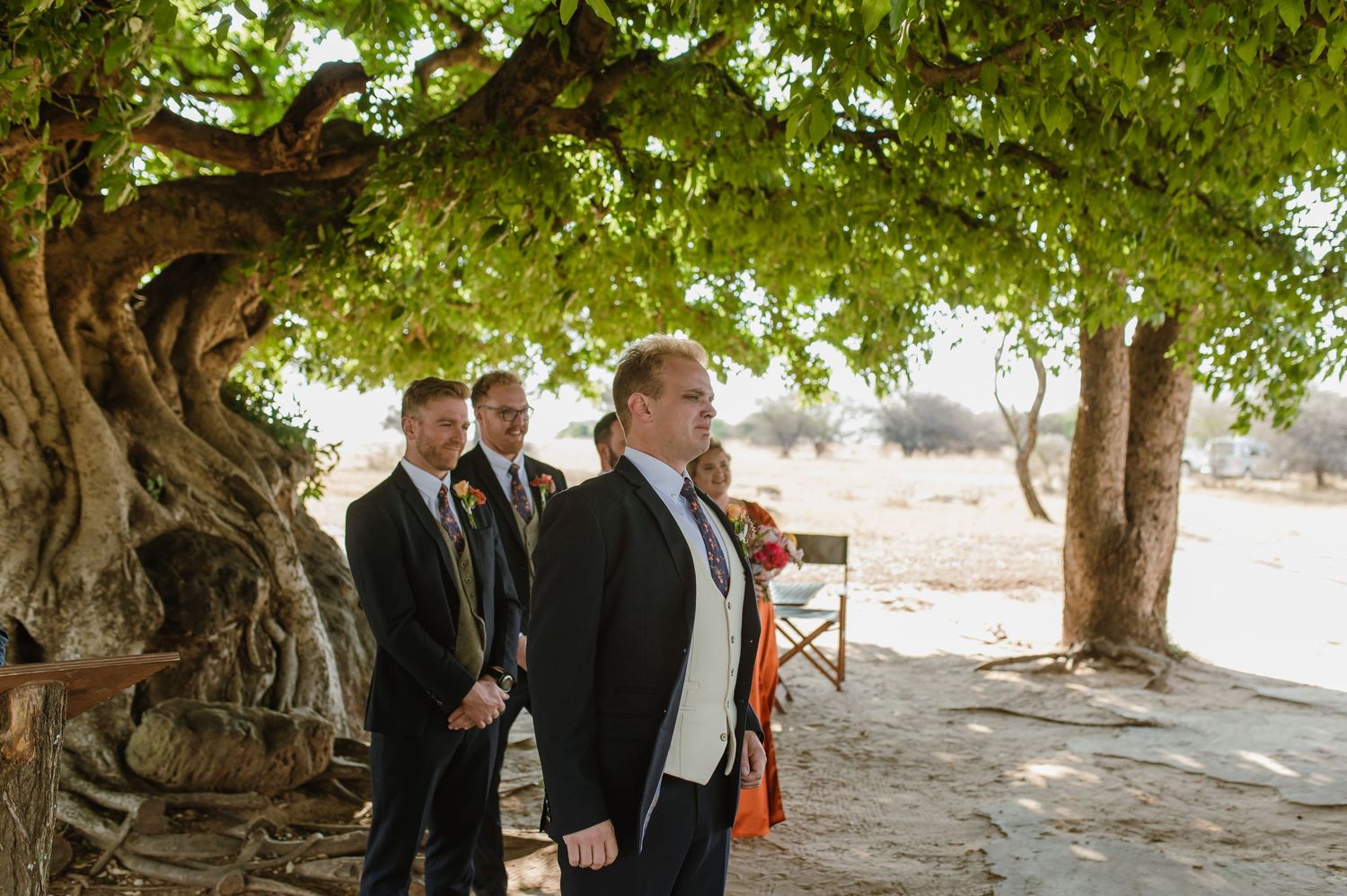 Guests gather under lush green trees for an outdoor wedding celebration.