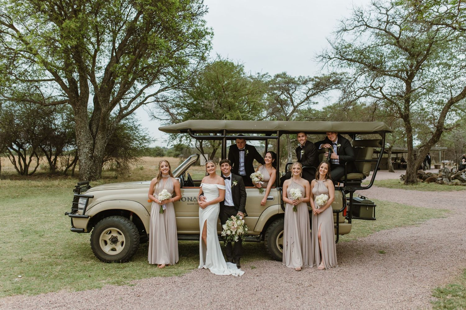 Wedding party gathered around classic cars on a dirt path surrounded by trees.