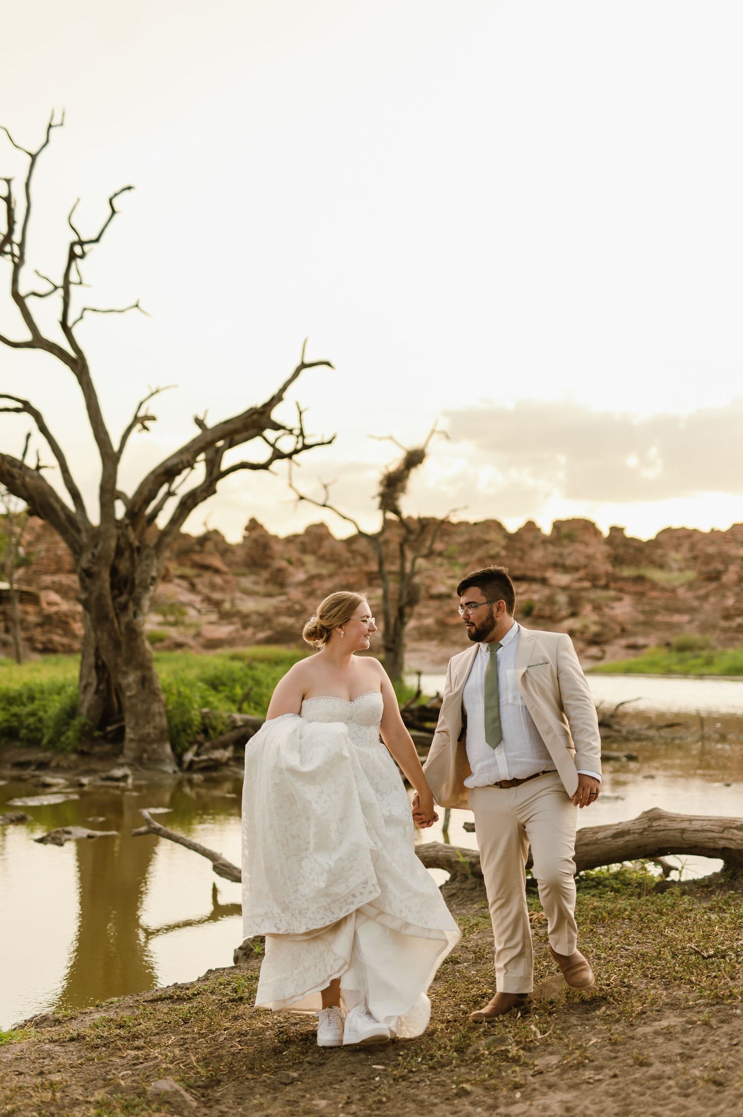 A couple in wedding attire shares a romantic moment by a scenic lake with bare trees at sunset.