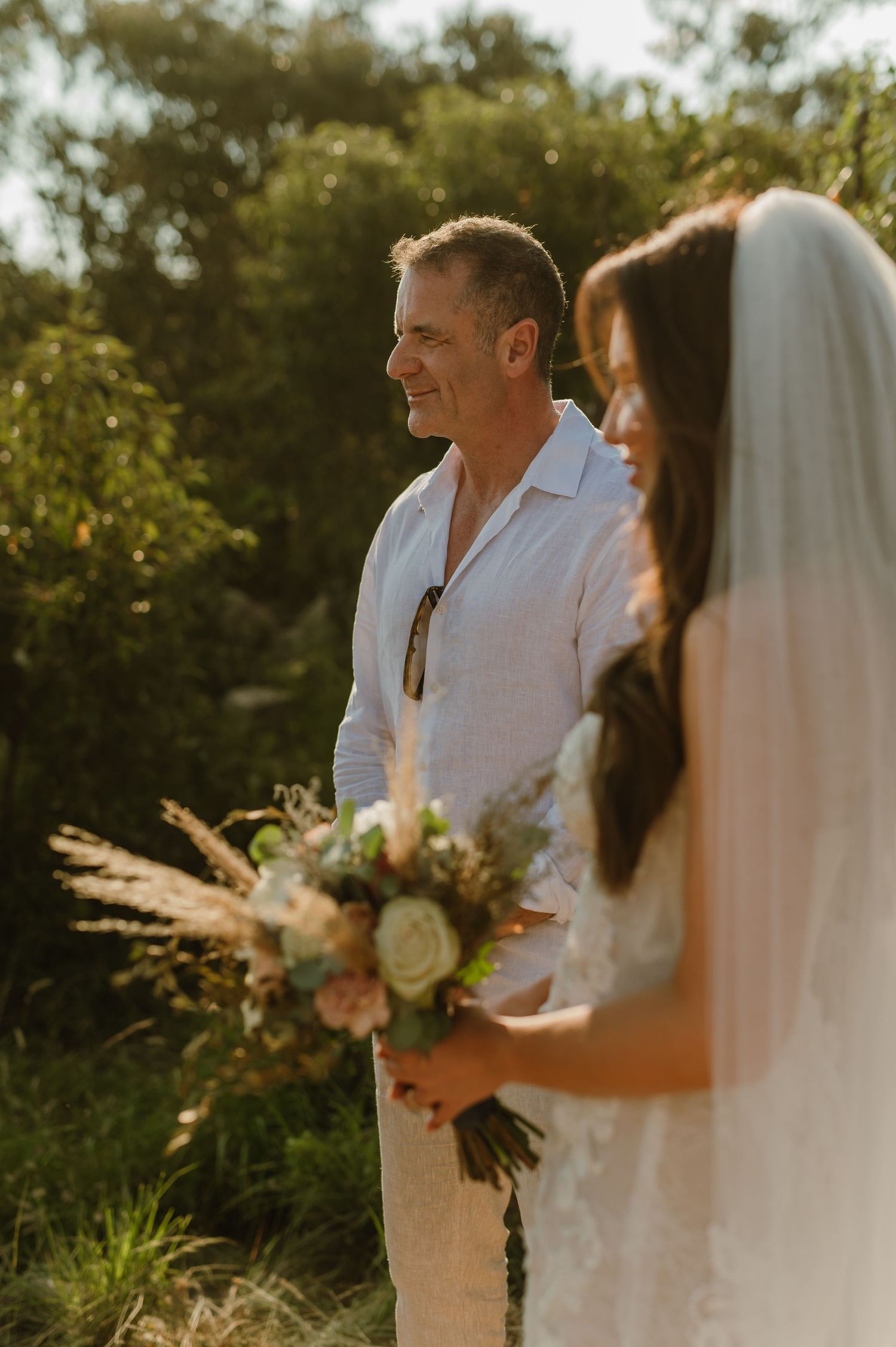 Groom smiling at bride holding rustic bouquet during golden hour outdoor safari bush wedding ceremony.