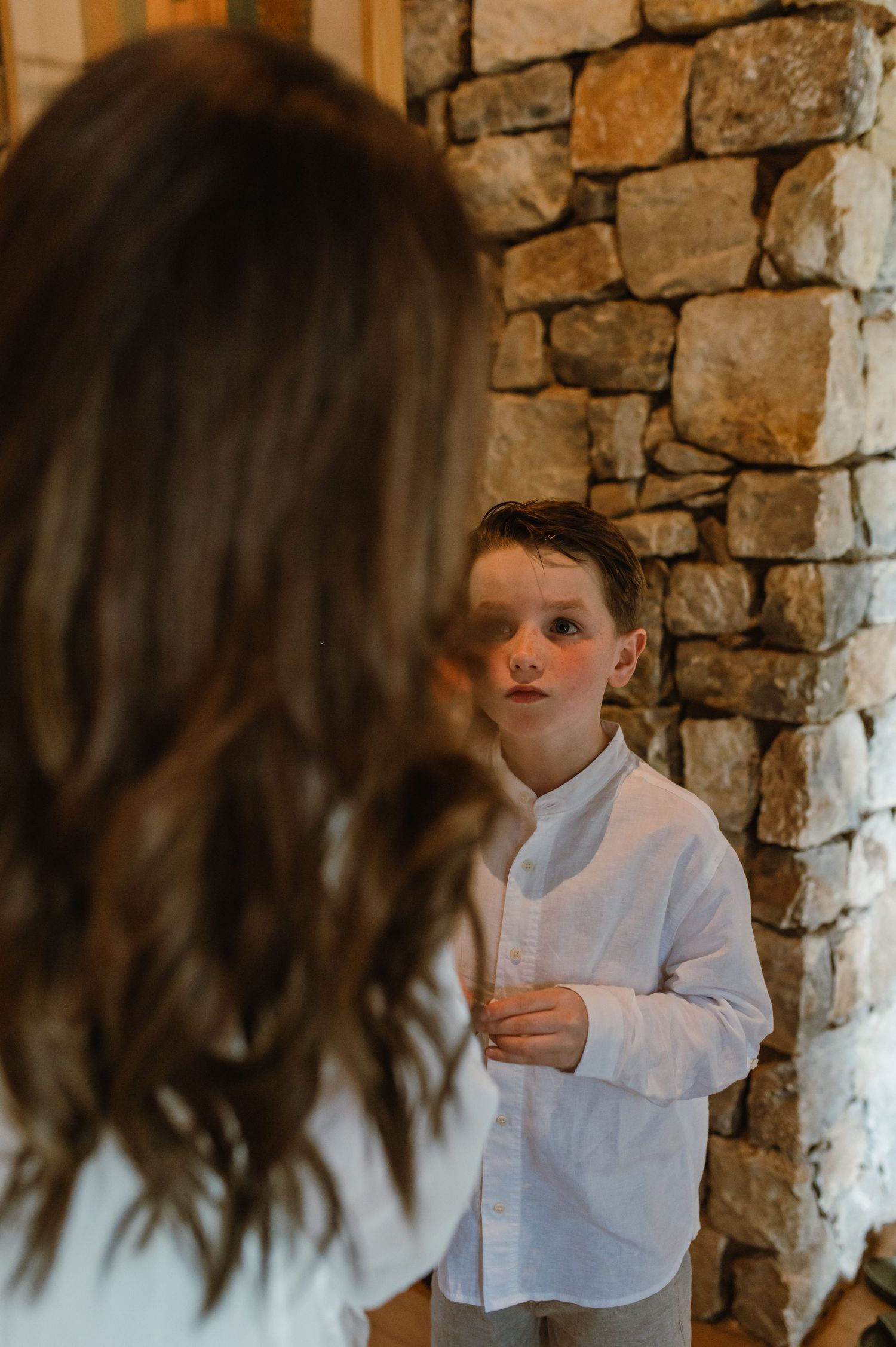 Young boy in white linen shirt standing against a stone wall at a wedding venue.