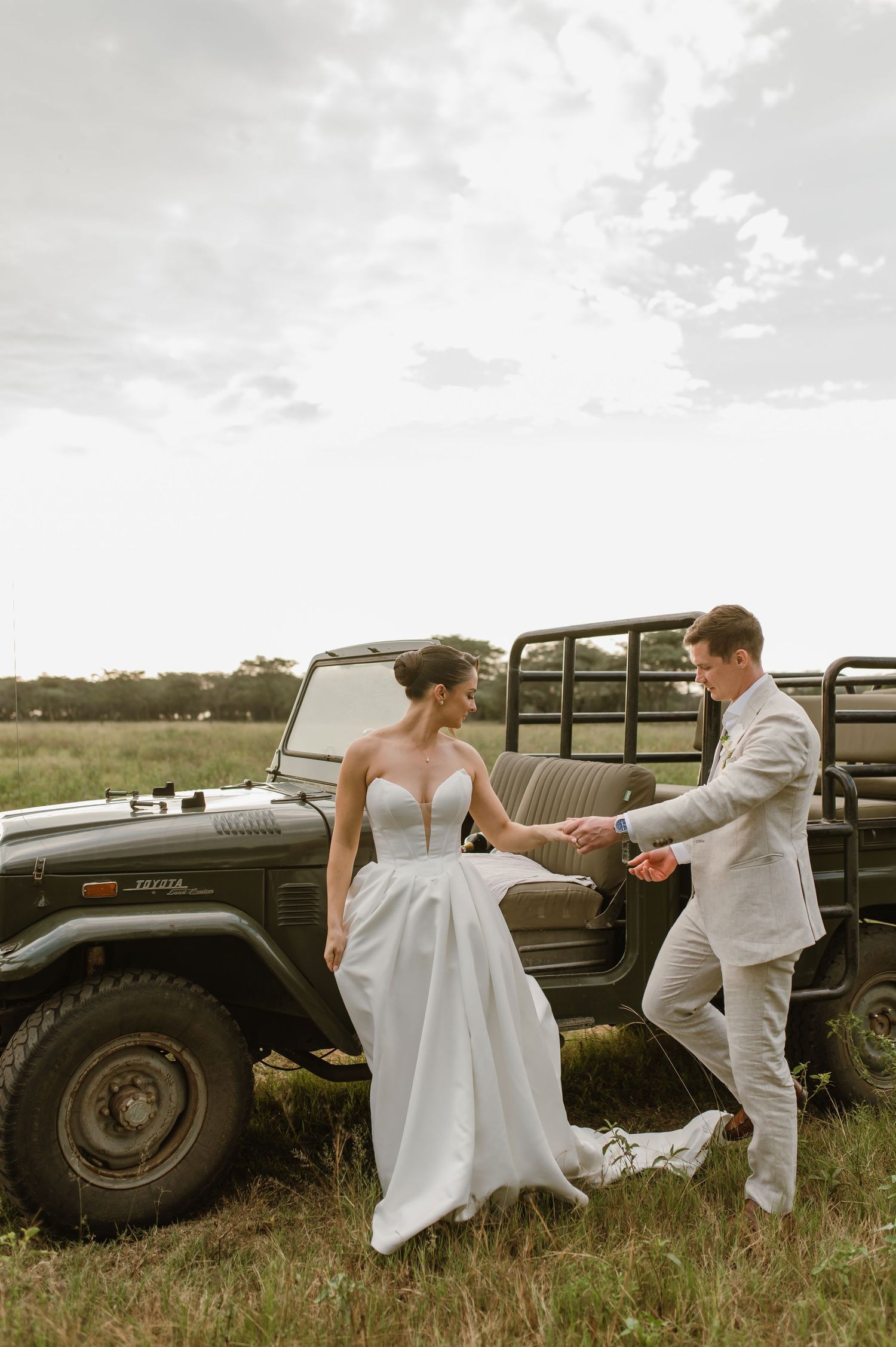 A couple in wedding attire playfully interacting near a vintage military vehicle in a grassy field.