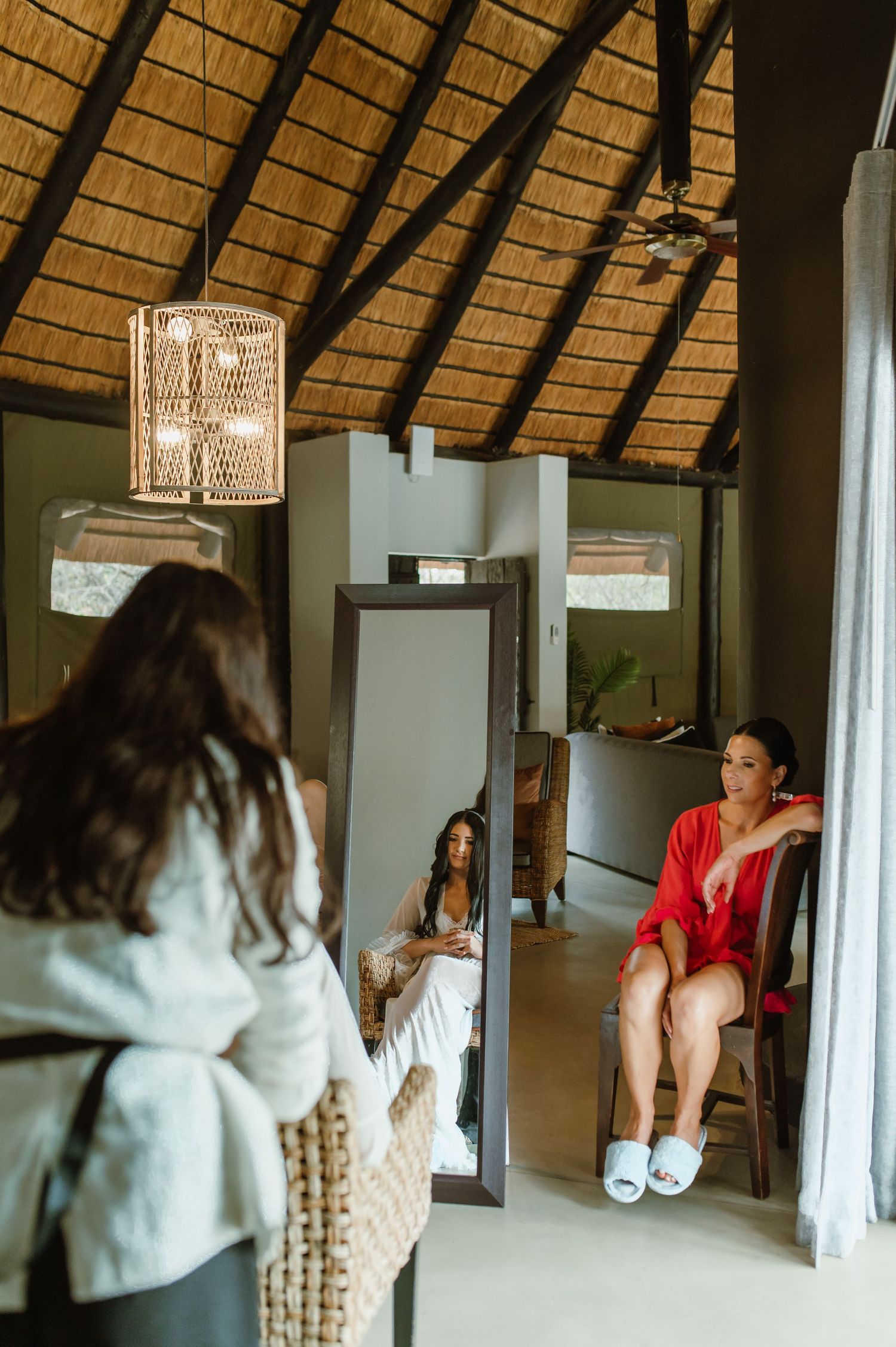 Bridesmaids getting ready in a thatched-roof safari lodge room with mirror and warm lighting.