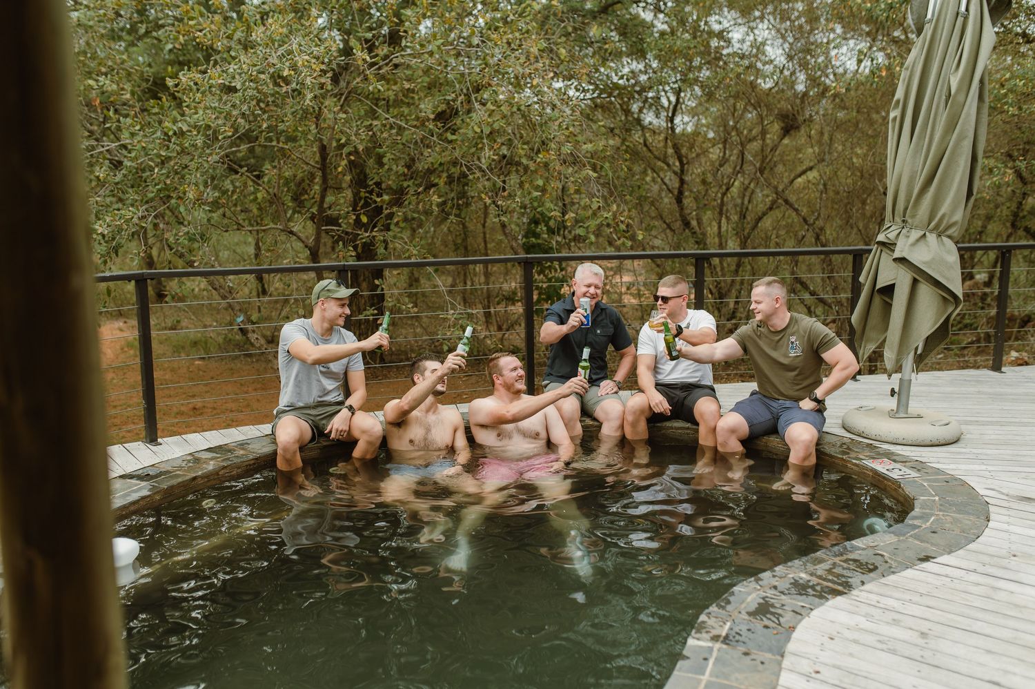 Group of men celebrating in an outdoor pool, raising bottles, surrounded by lush trees on a wooden deck.