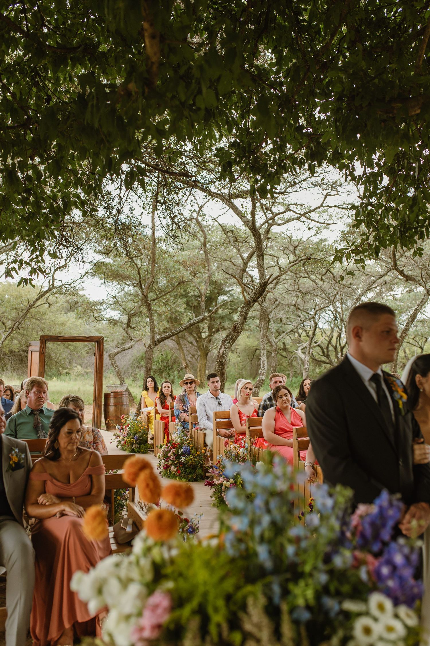 Outdoor wedding ceremony under lush trees with colorful floral arrangements and seated guests.