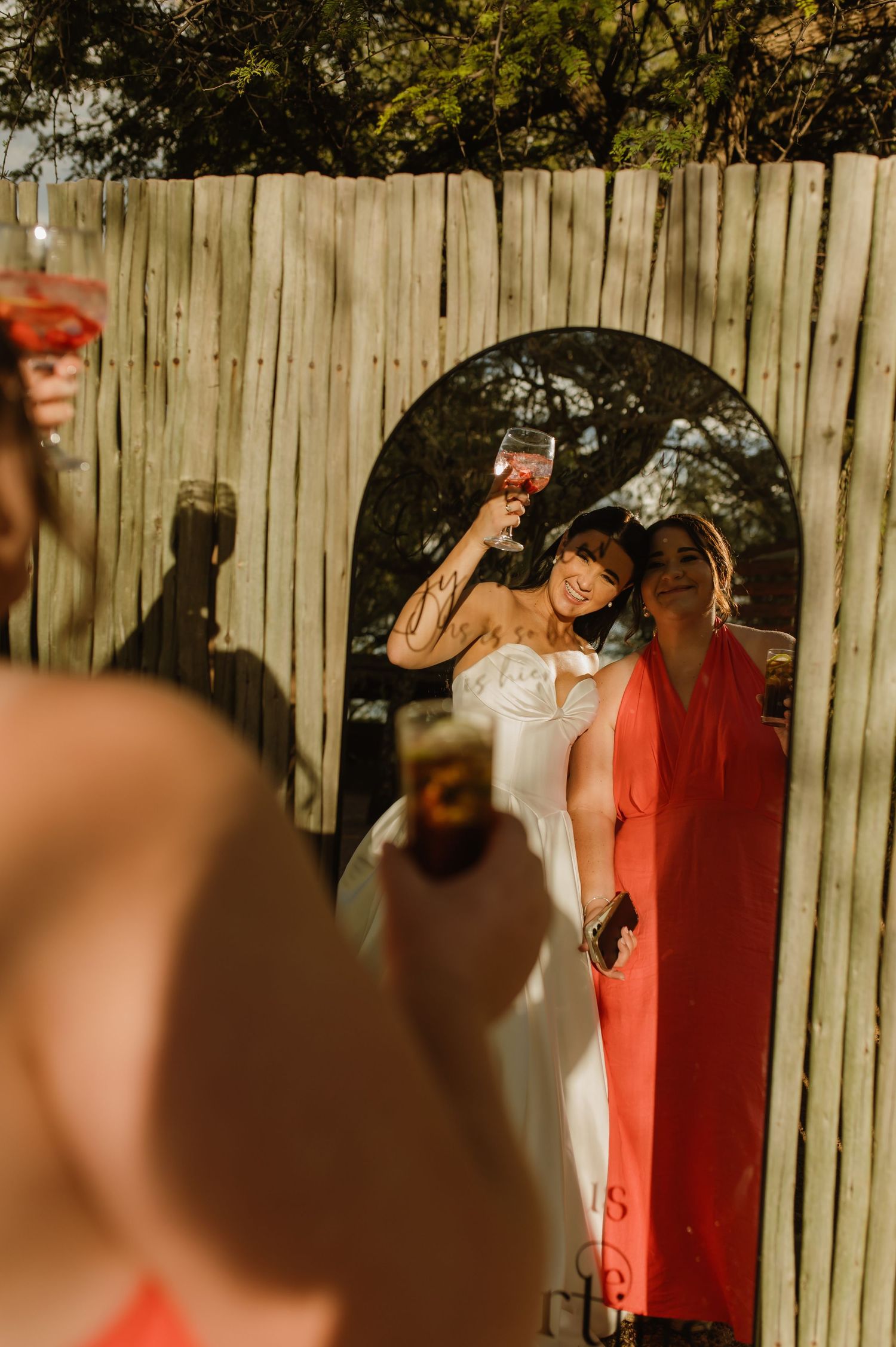 Bride in white strapless gown raises wine glass with bridesmaid in red dress at rustic wooden fence backdrop.