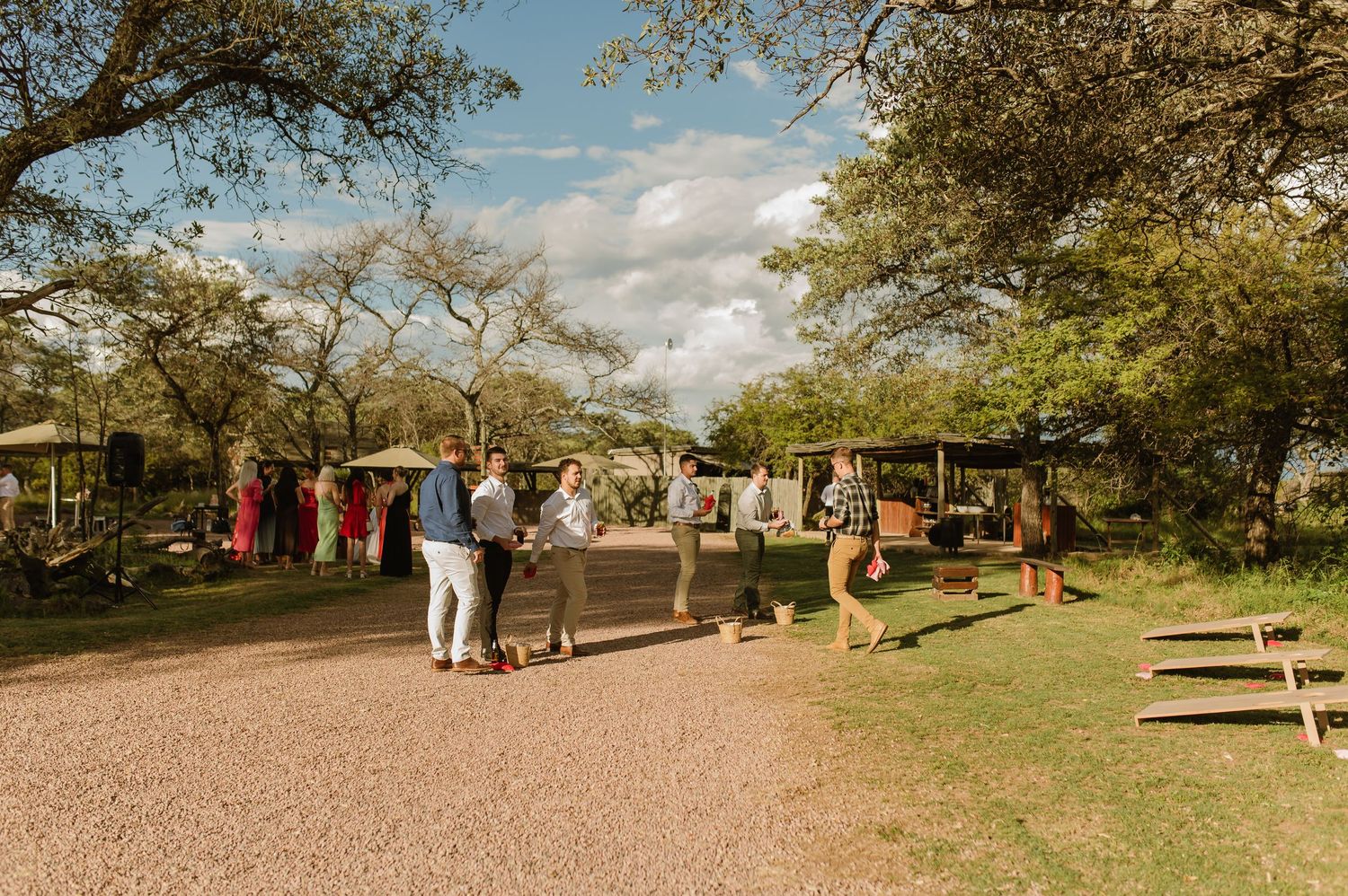 Wedding guests mingle outdoors under trees at rustic open-air reception venue on a sunny day.