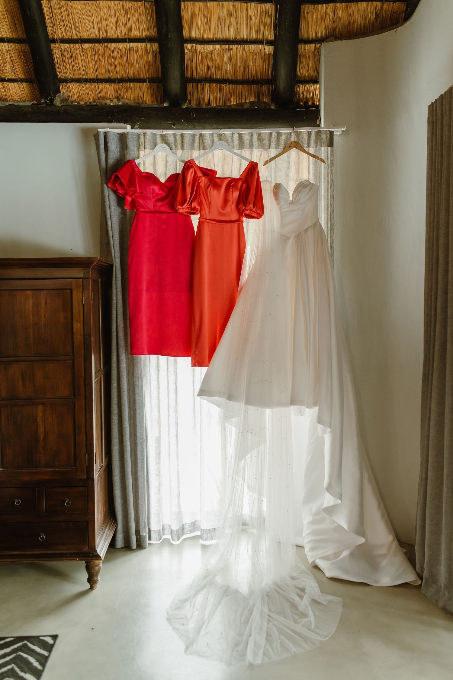 Wedding dress and red bridesmaid dresses hanging on a curtain rod in a rustic thatched-roof room.