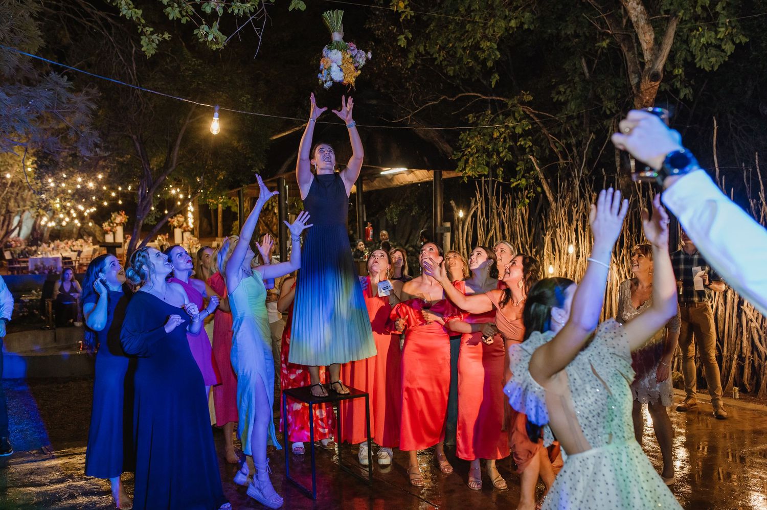 Bride lifted above crowd during bouquet toss at a vibrant outdoor nighttime wedding reception under string lights.