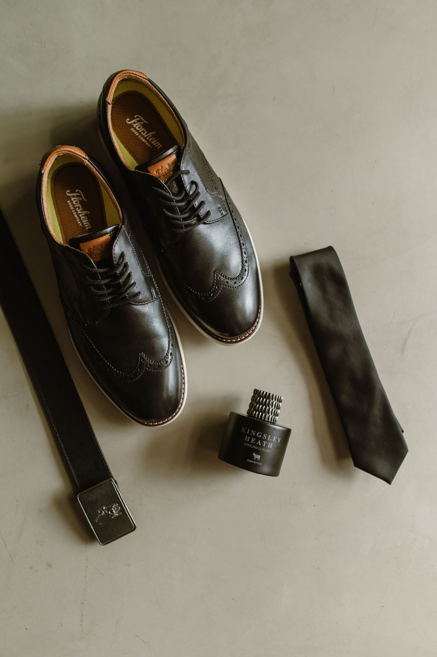 Dark brown wingtip brogue shoes with belt, tie, and cologne arranged on gray surface for groom wedding day details.