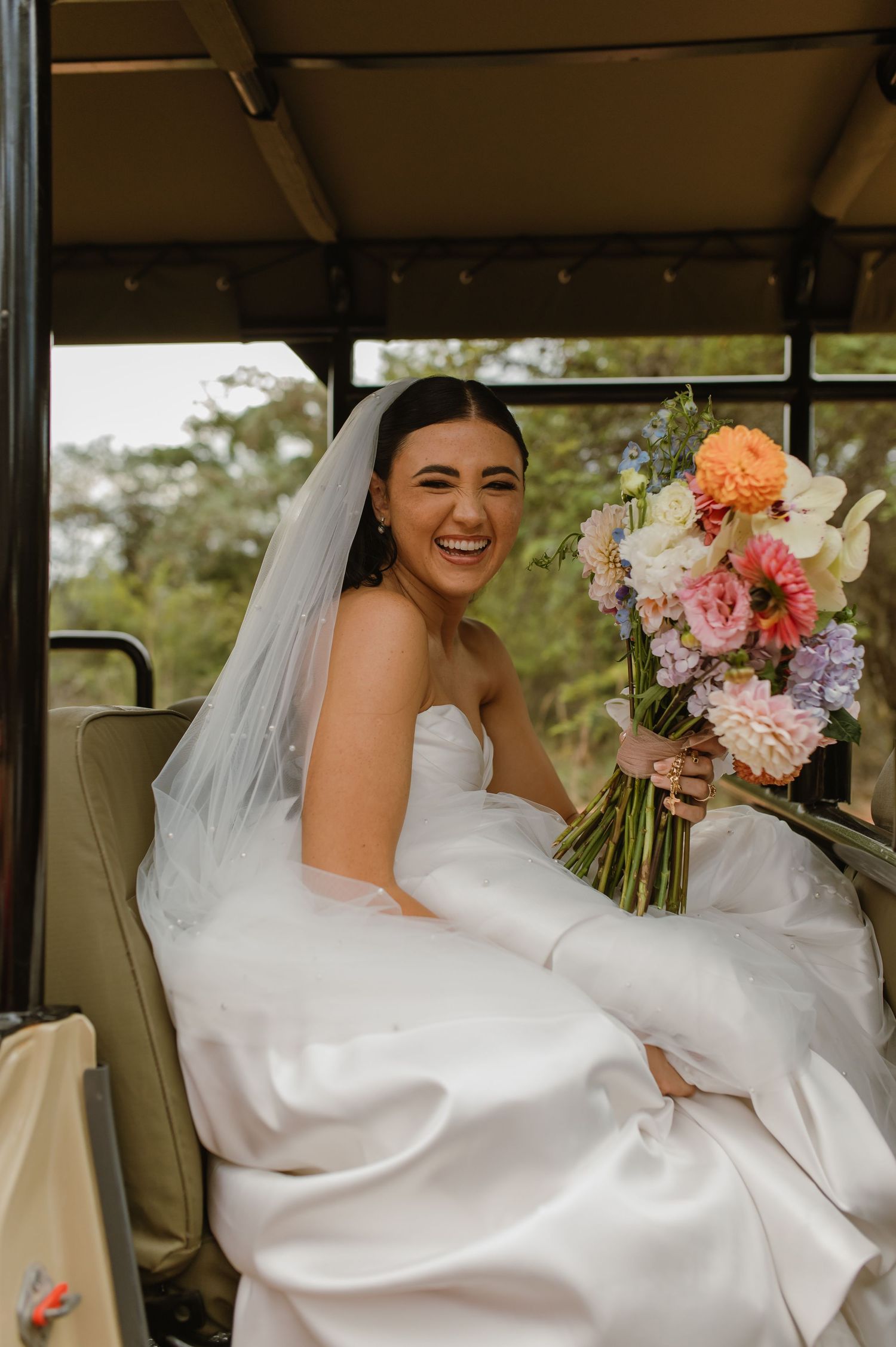 Laughing bride in white strapless gown and veil holding a colorful bouquet while seated in a safari vehicle.