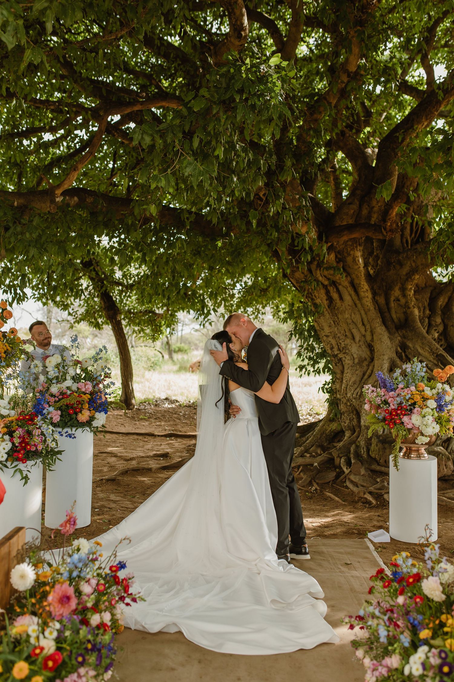 Bride and groom sharing first kiss under a large tree surrounded by vibrant floral arrangements.