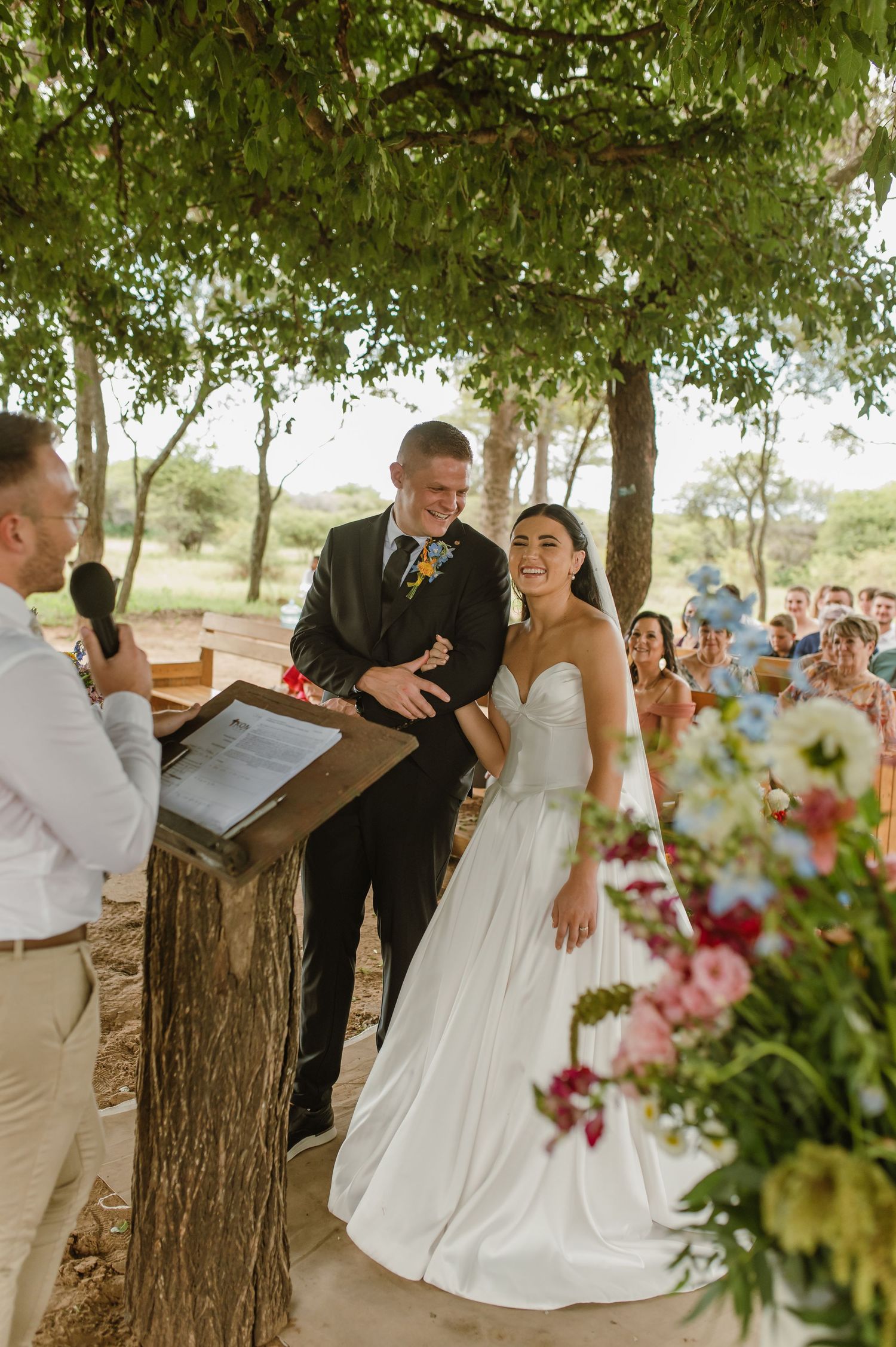 Smiling bride and groom exchange vows at outdoor wedding ceremony surrounded by guests and floral arrangements.