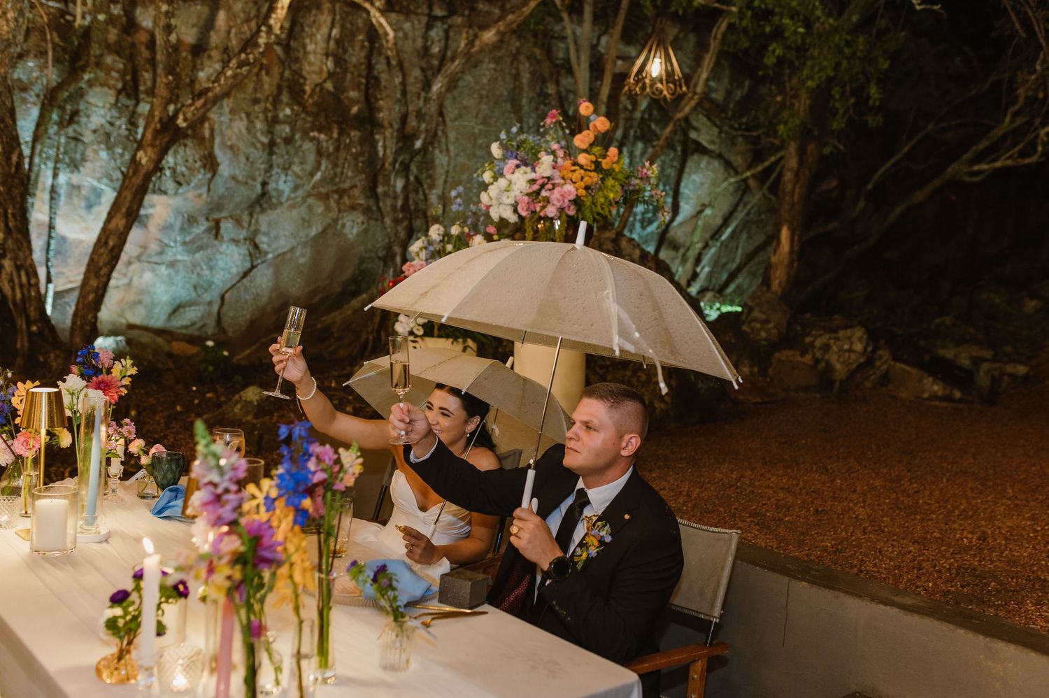 Groom taking selfie under umbrella at wedding sweetheart table adorned with colorful flowers and gold candleholders.