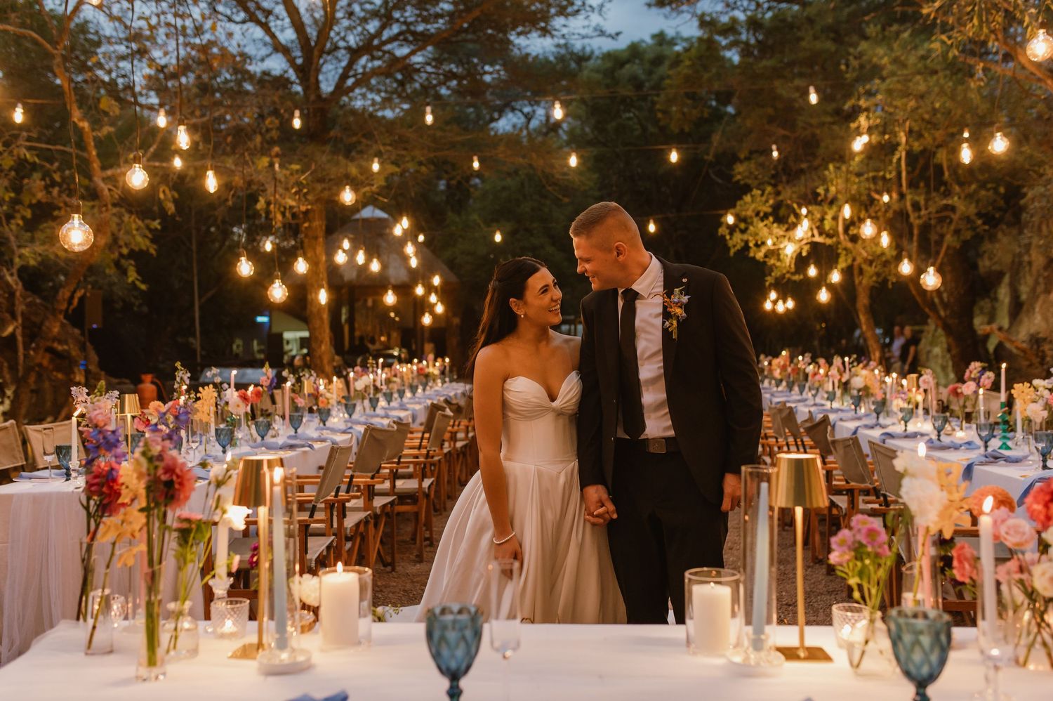 Newlyweds share a romantic moment at their elegantly lit outdoor reception with string lights and floral decor.