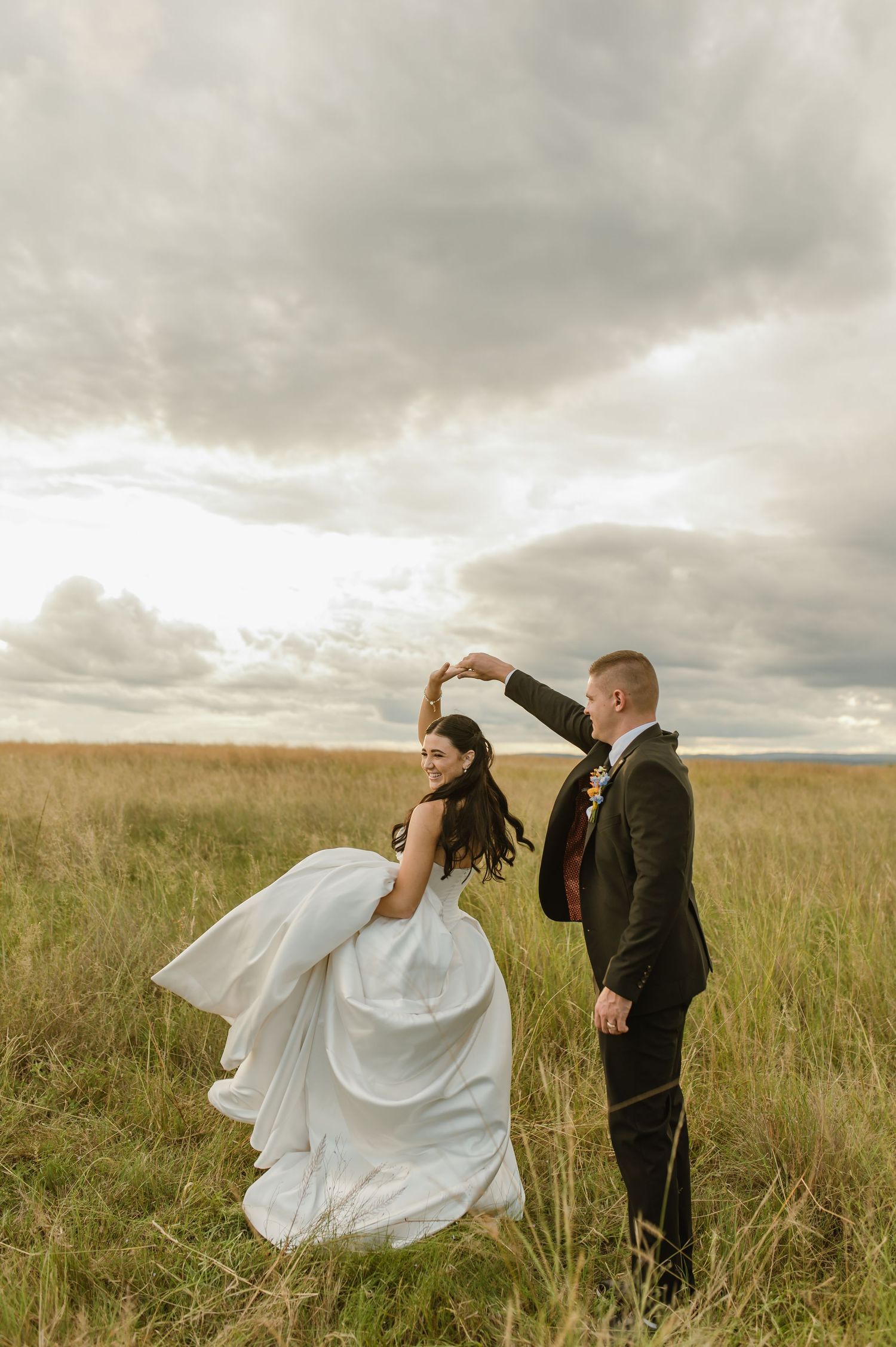 Bride and groom dancing in a golden grass field under dramatic cloudy skies at sunset.