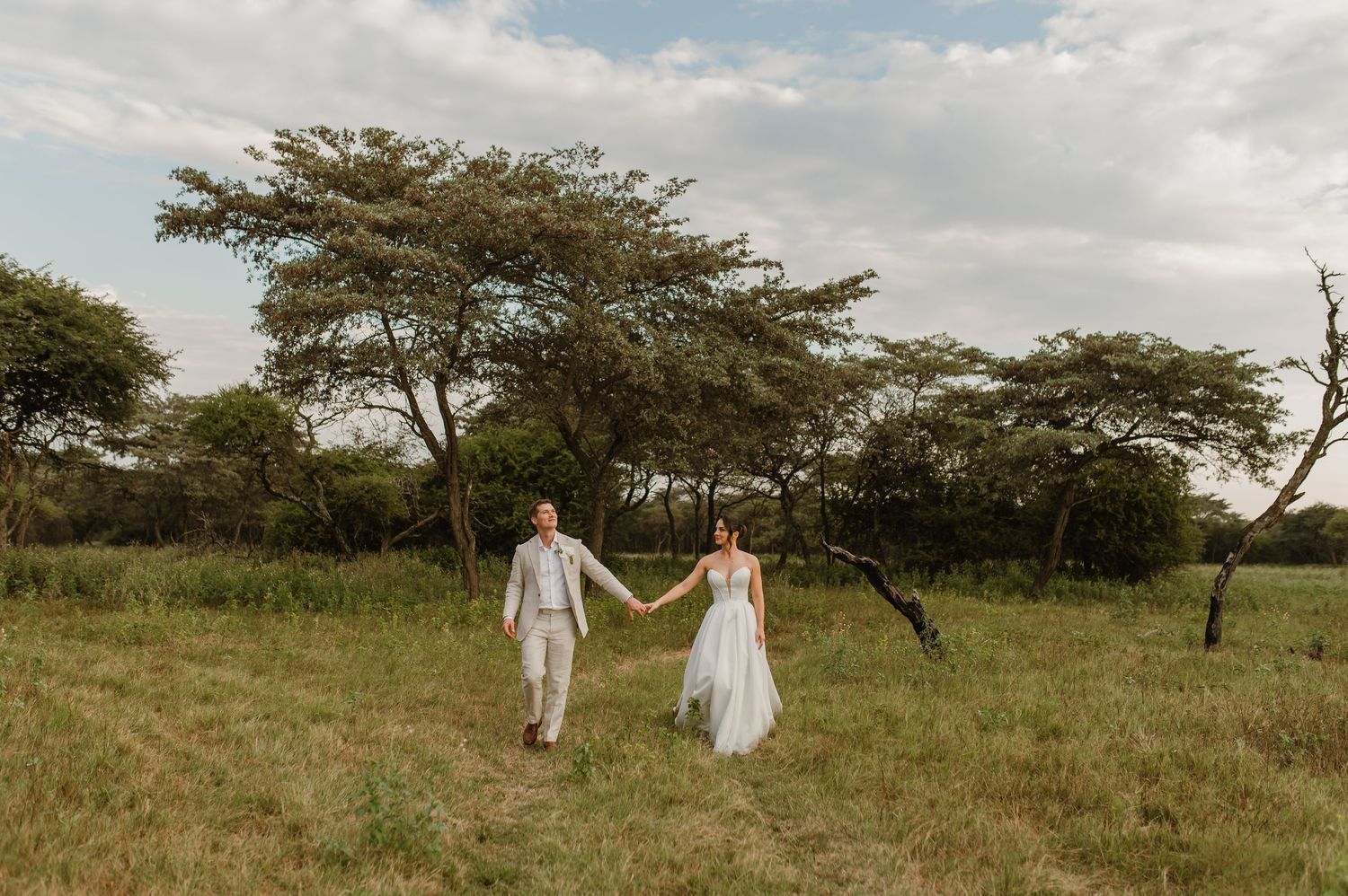 A romantic wedding photo shoot in a vast African savanna with scattered acacia trees under cloudy skies.
