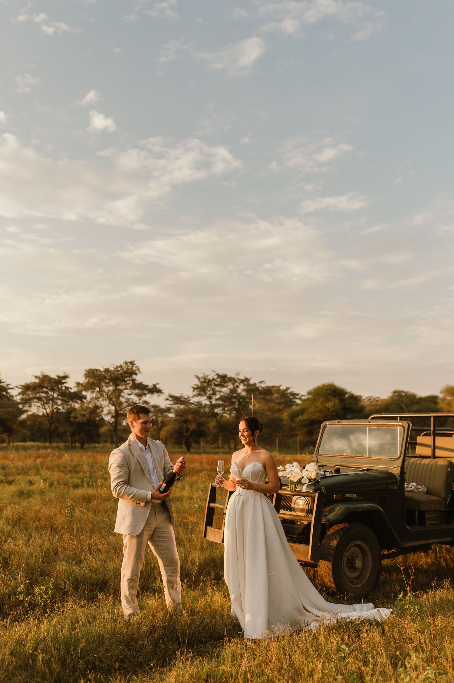 A wedding couple poses next to a vintage vehicle in an African safari setting during golden hour.