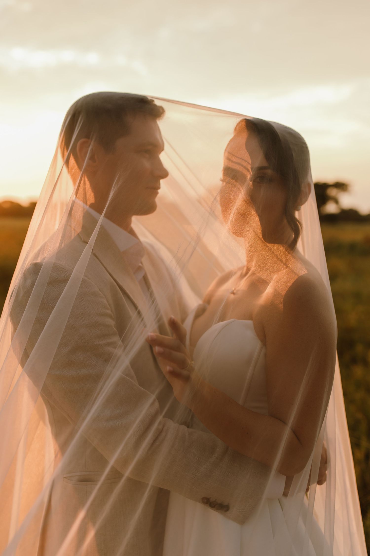 A romantic wedding couple embraces under a flowing veil at sunset in a field.