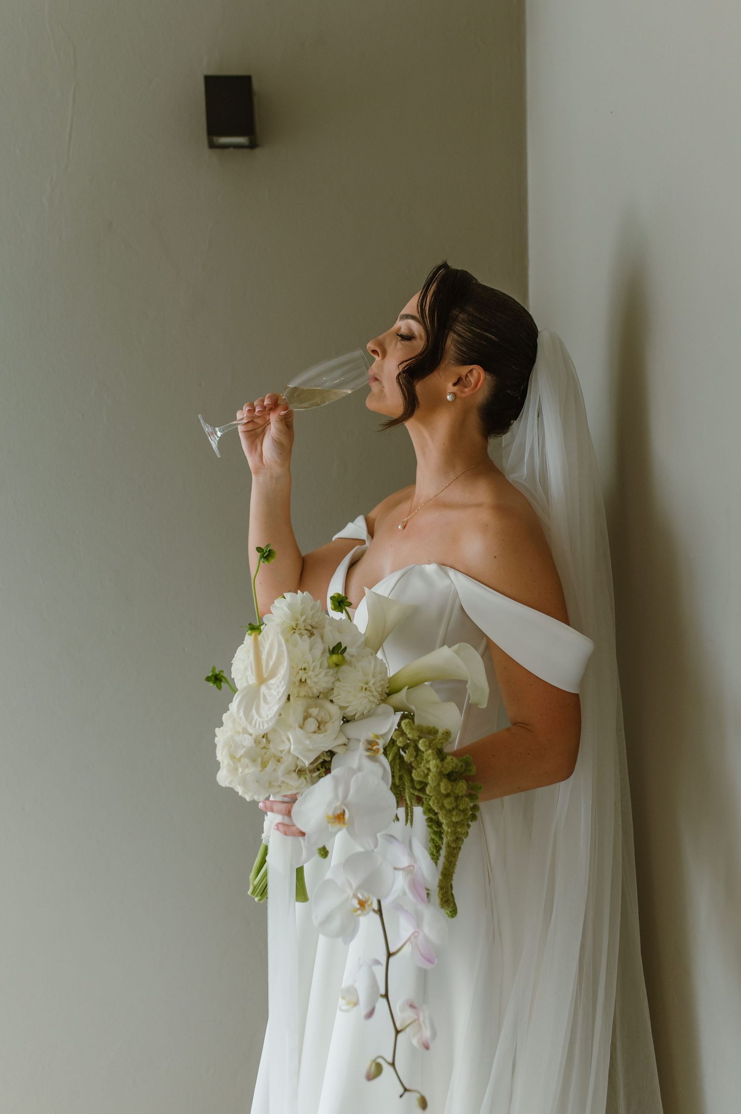 Person in white wedding dress holding bouquet while drinking from champagne bottle.