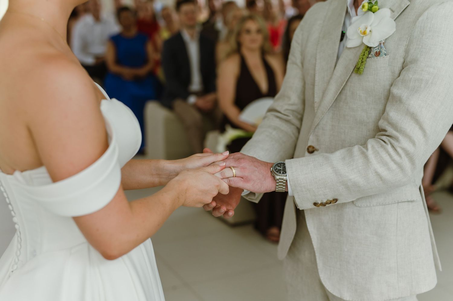 A close-up shot of a couple's hands during a wedding ring exchange ceremony in a bright indoor setting.