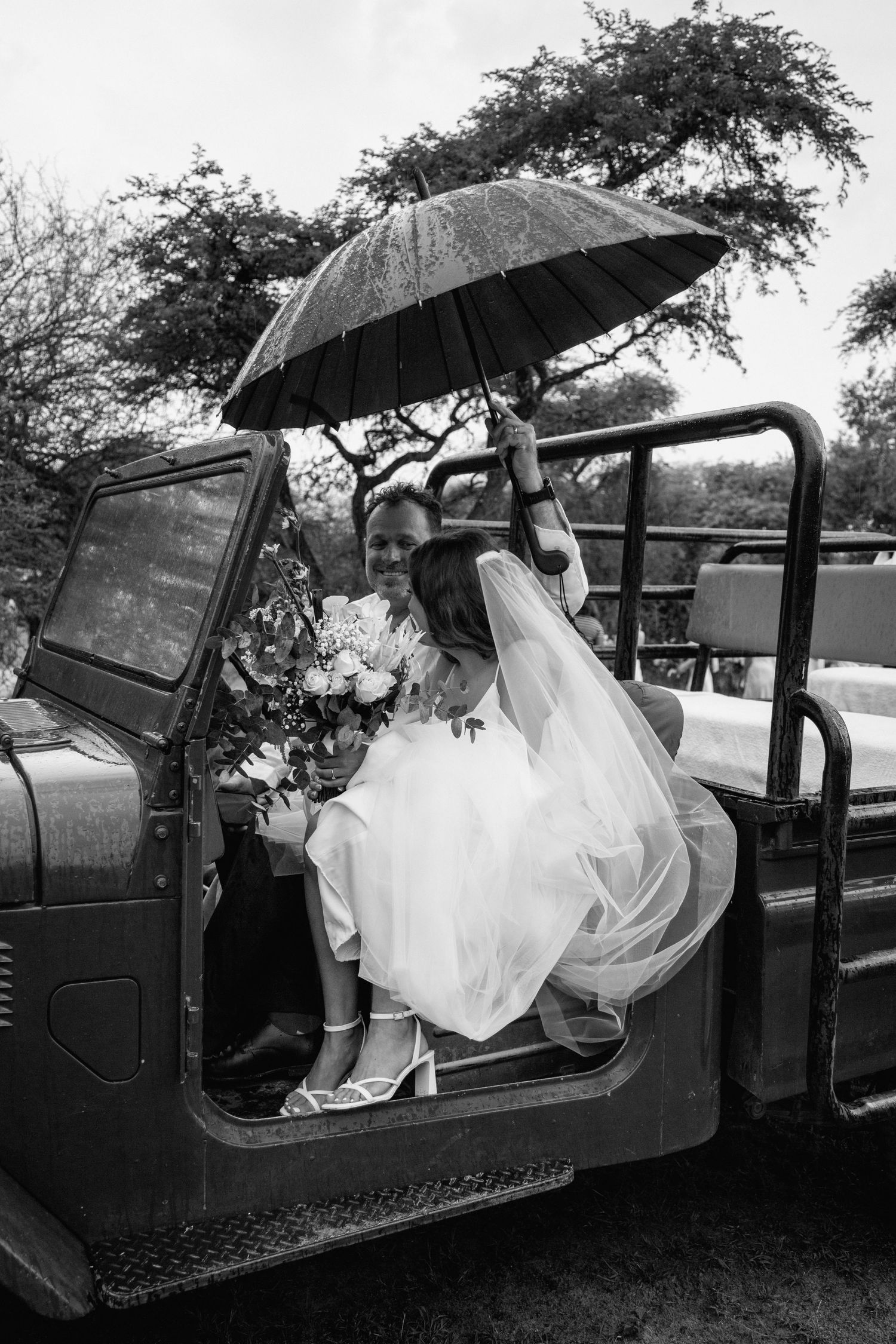 Black and white photo of couple in vintage vehicle under umbrellas at wedding celebration.