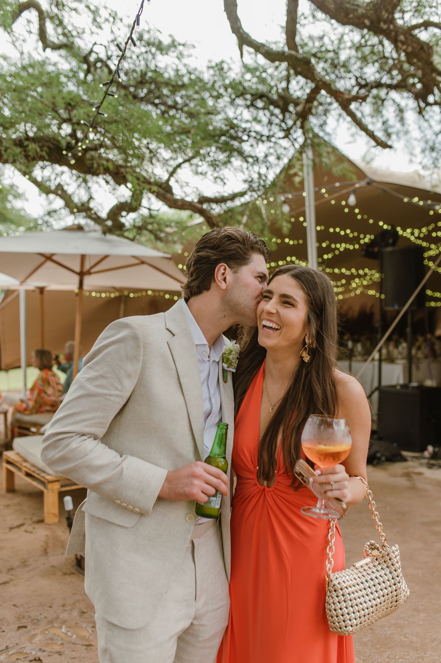 Couple sharing drinks and laughter at outdoor wedding reception under string lights.