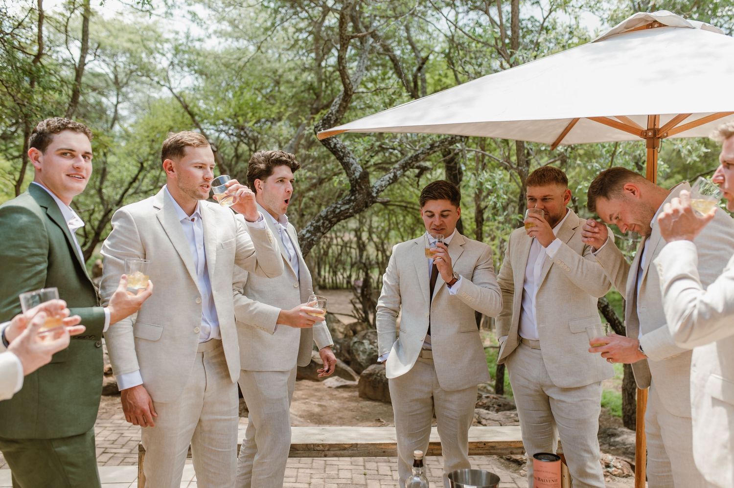 Wedding party members in coordinated beige suits toast together in a natural outdoor setting.