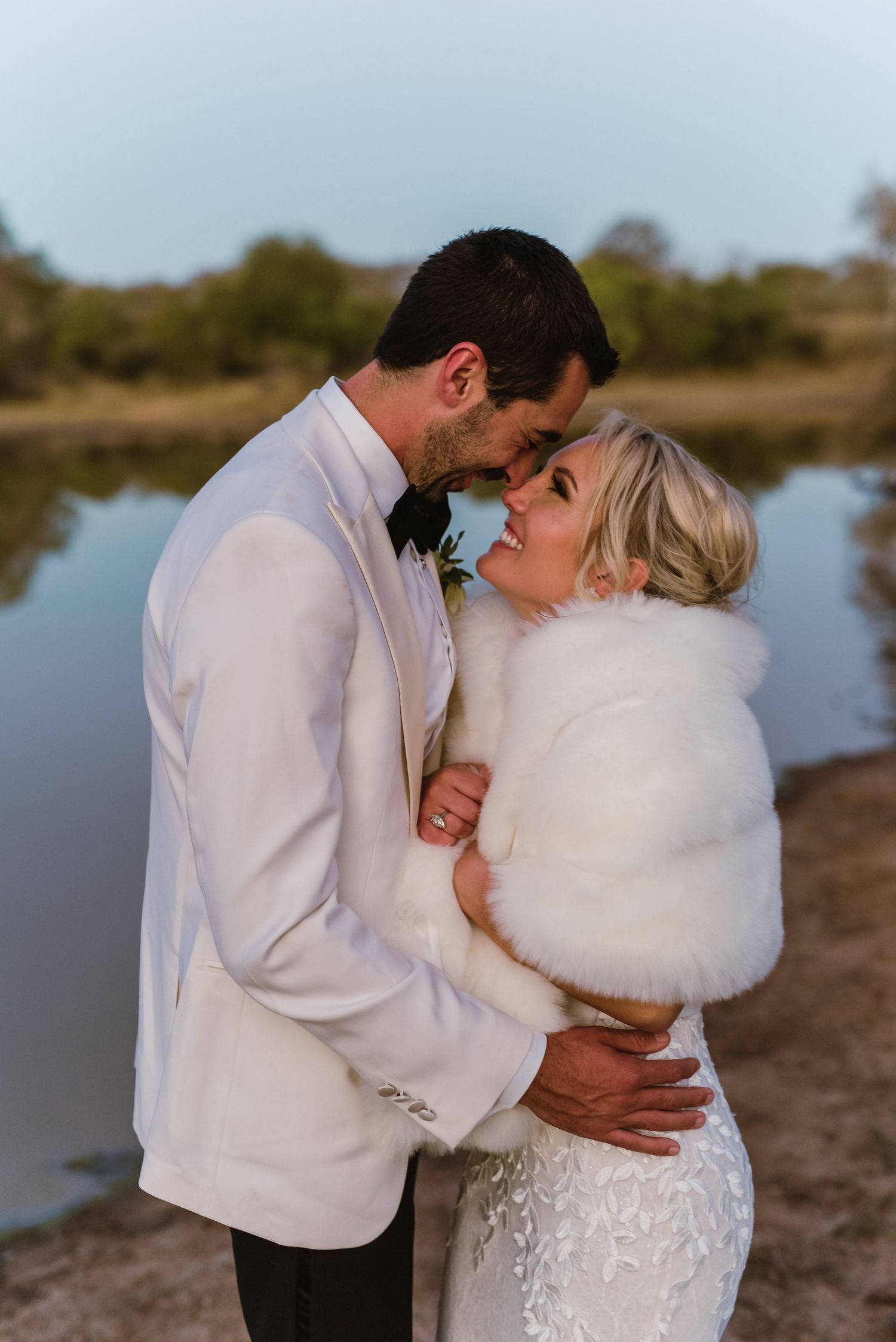 A romantic moment between newlyweds in white attire by a serene waterfront.