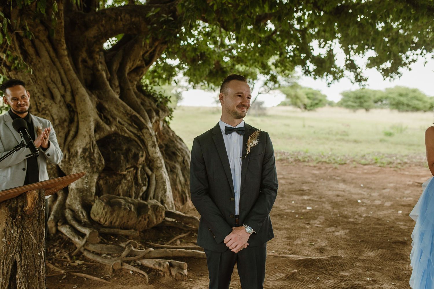 Wedding ceremony guests gather under a canopy of trees in an outdoor rustic setting.
