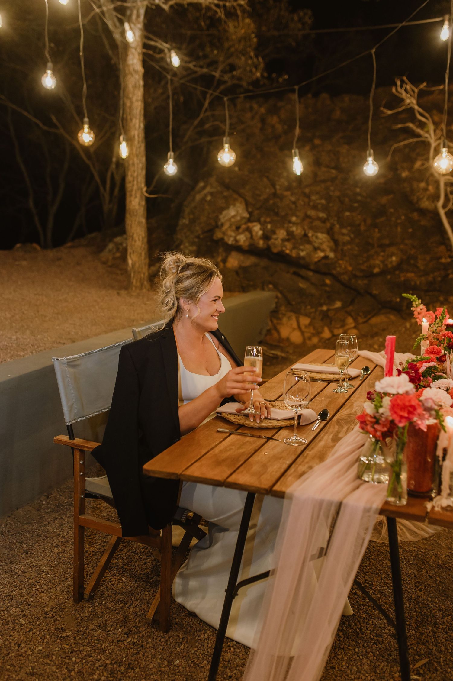 Guest seated at wooden table with string lights and floral decorations at outdoor evening event.