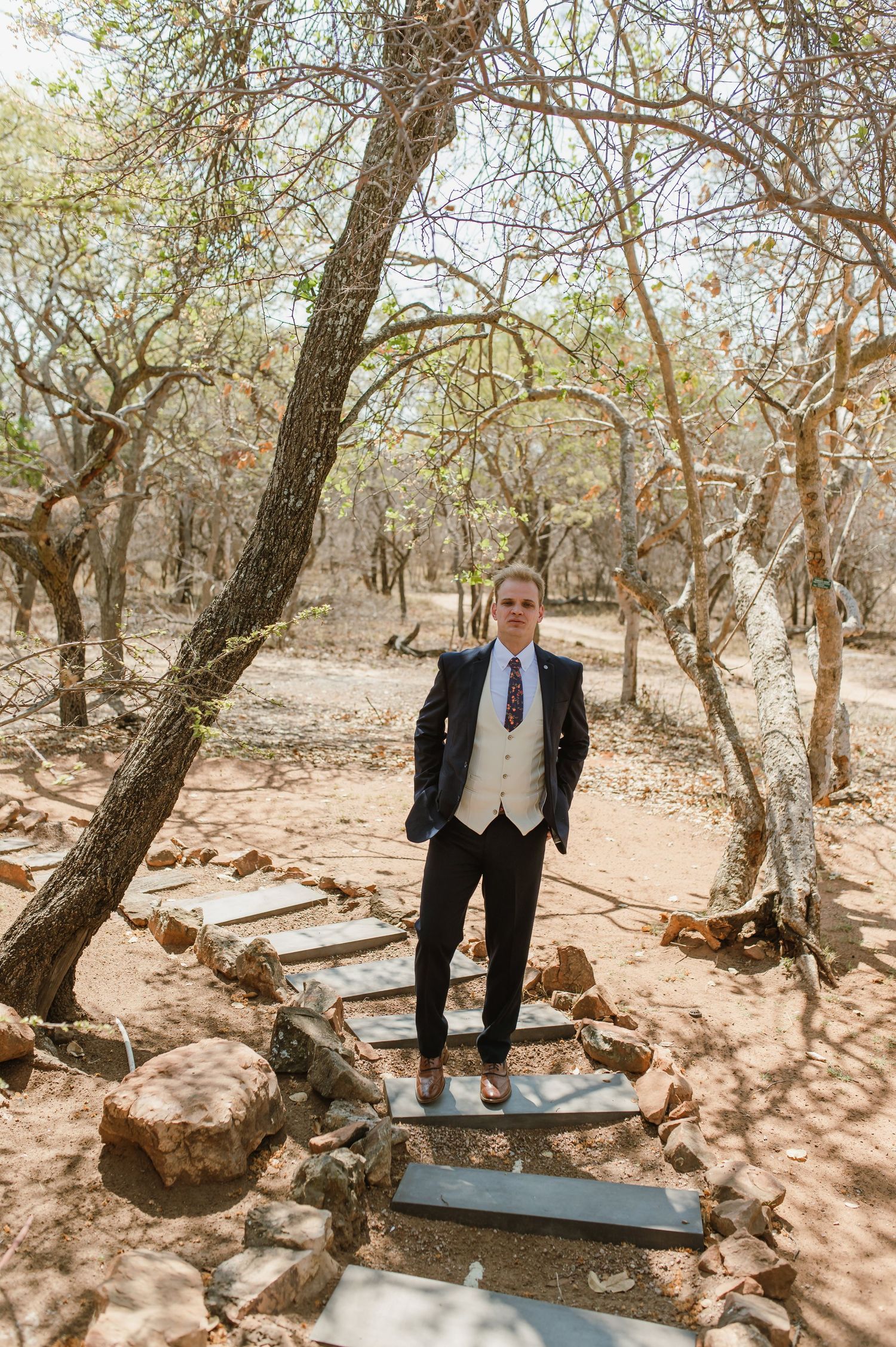 A formal suited figure stands on stone steps among bare trees in a natural outdoor setting.