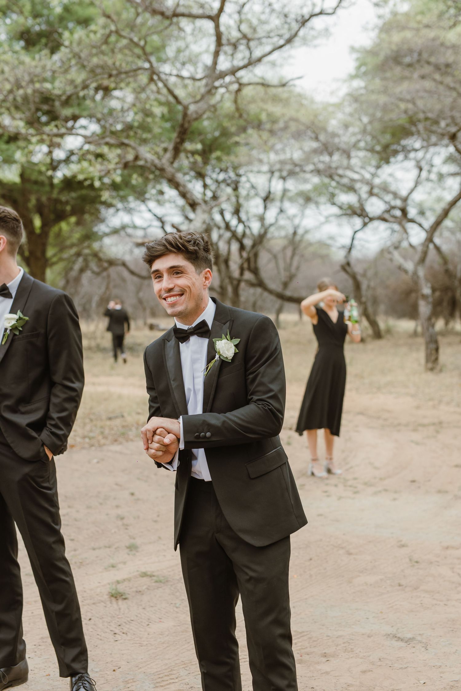 Groomsman in black tuxedo laughing during outdoor wedding festivities.