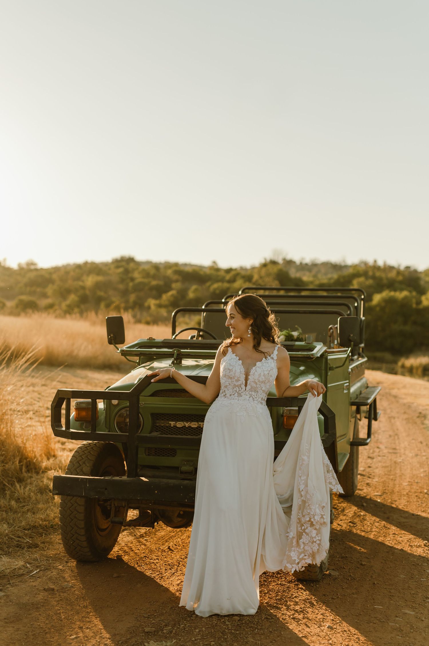 Two people in white dresses pose next to a black off-road vehicle during sunset.