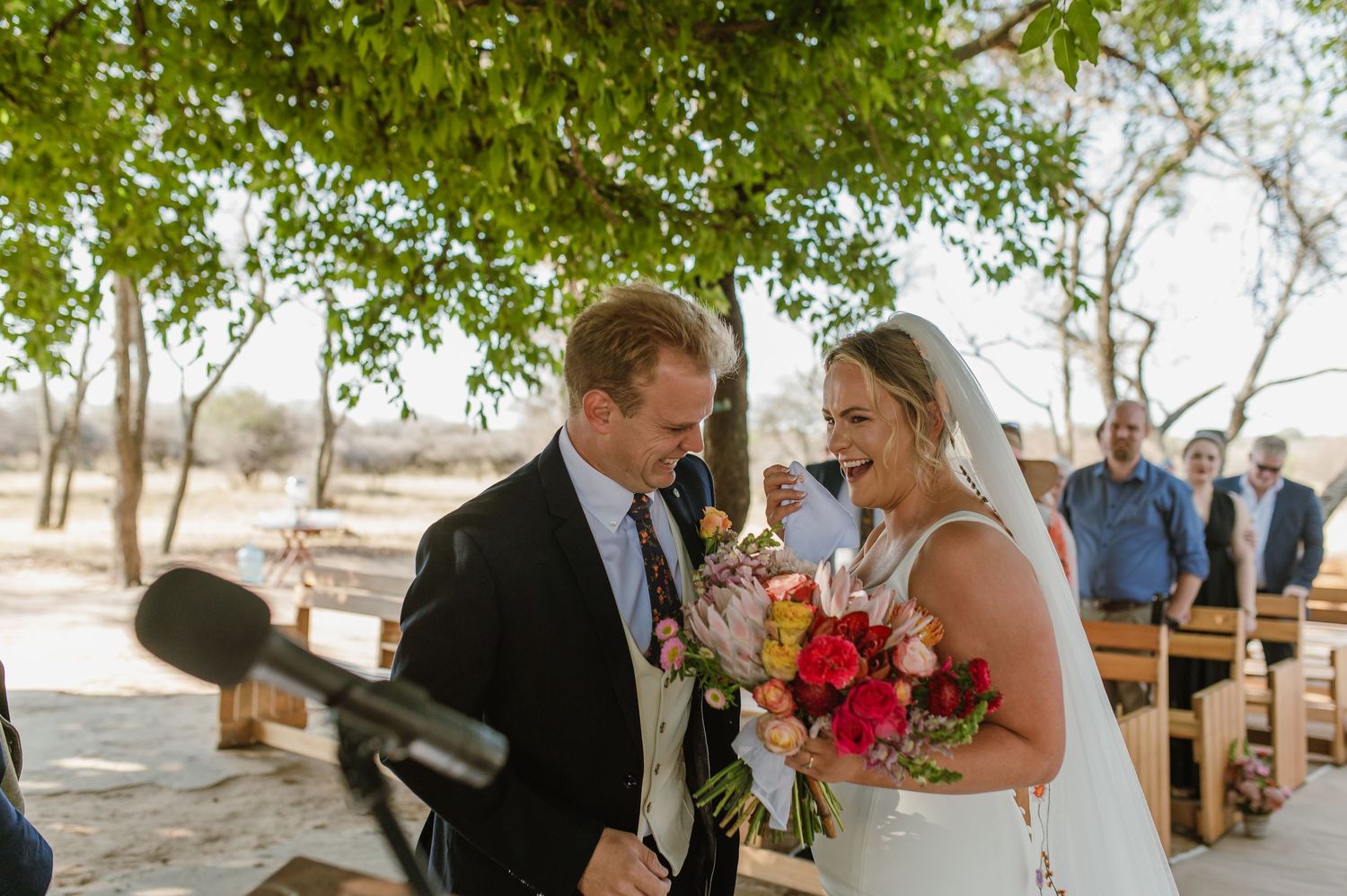 A romantic outdoor wedding ceremony takes place under a canopy of green trees.